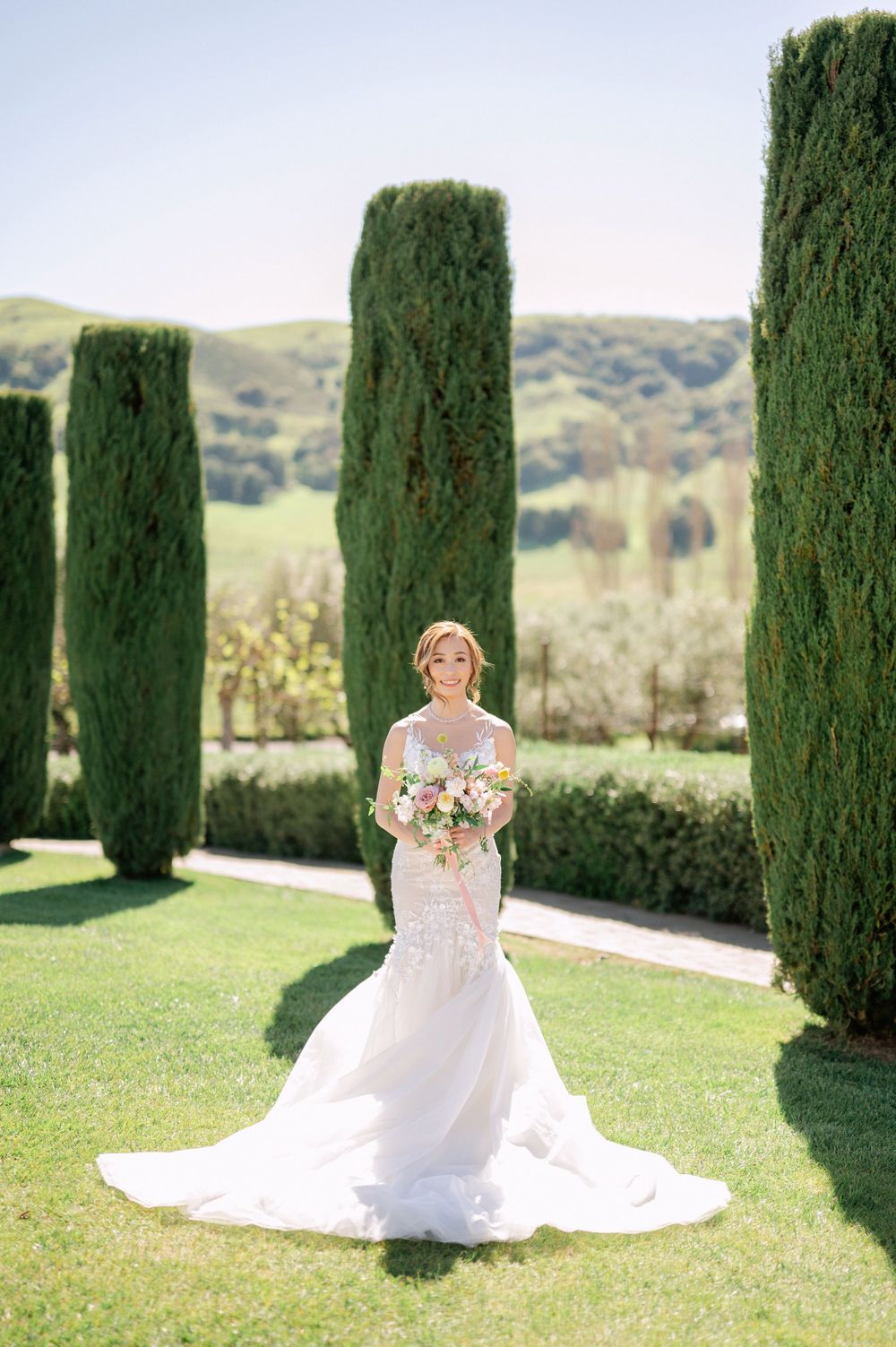 Bride in white lace gown stands alone amidst tall cypress trees on manicured lawn.