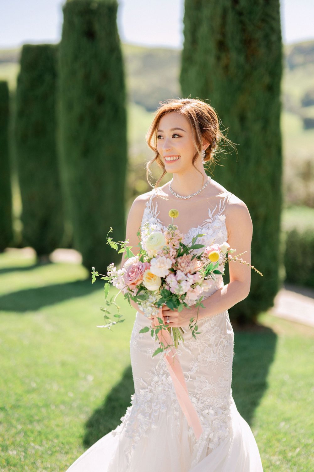 Bride holds spring bouquet of pink and white flowers while smiling in sunlit garden.