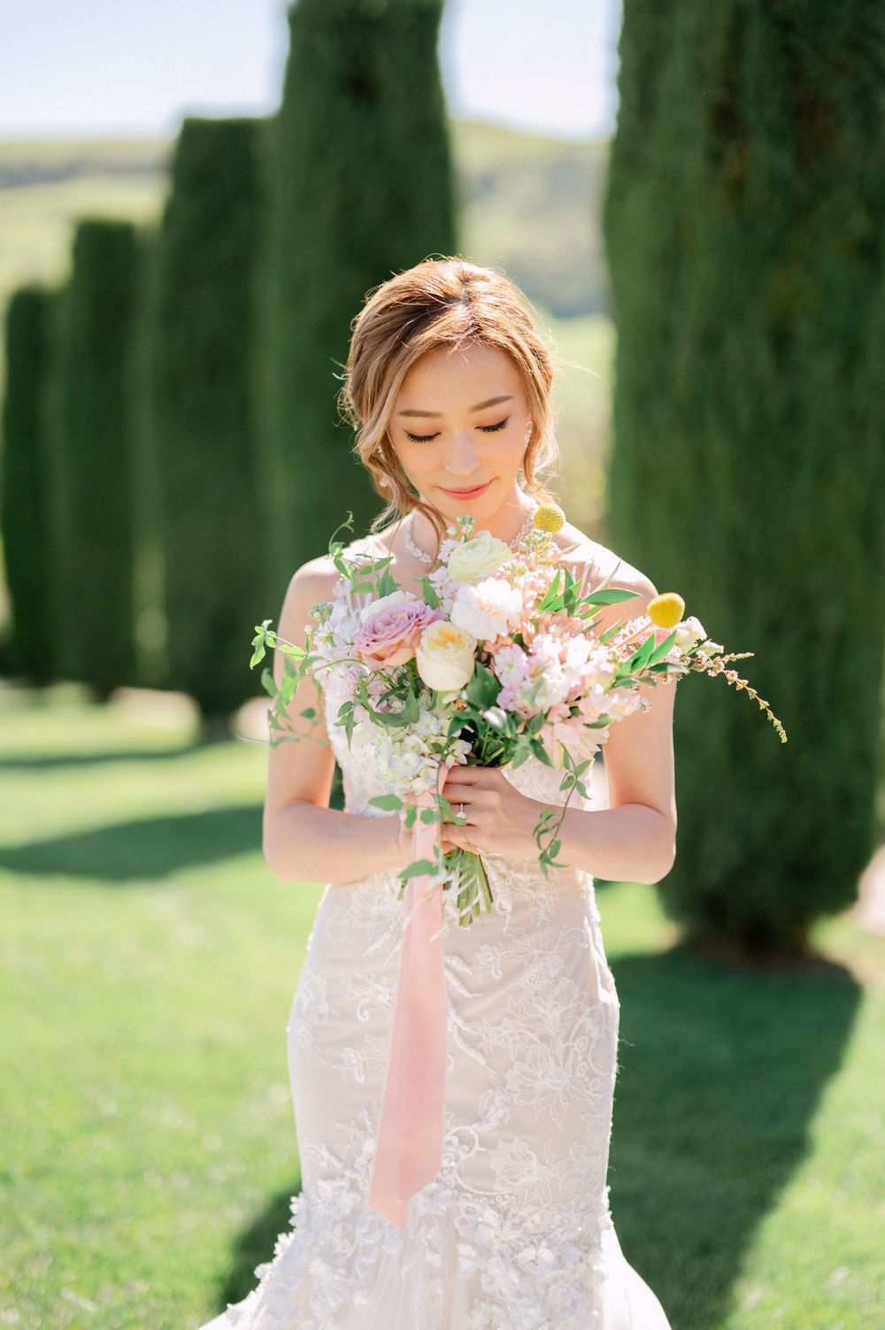 Bride gazes down at delicate pastel bouquet against backdrop of cypress trees.