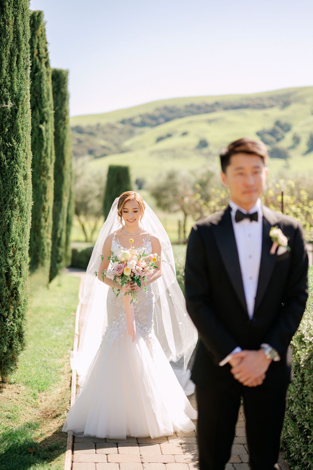 First look wedding photo with bride and groom on stone path lined with cypress trees.
