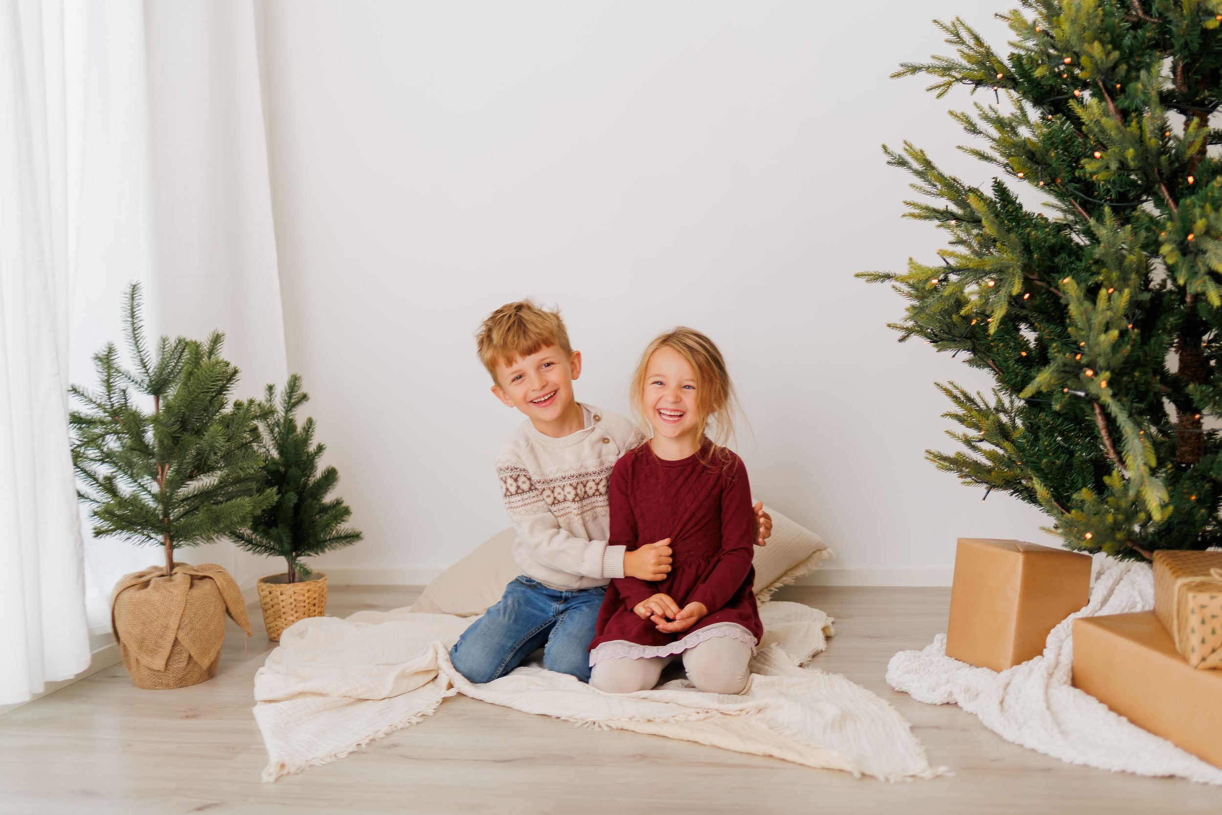 Two happy children sit together on a blanket surrounded by Christmas trees and gift boxes in a bright white room.