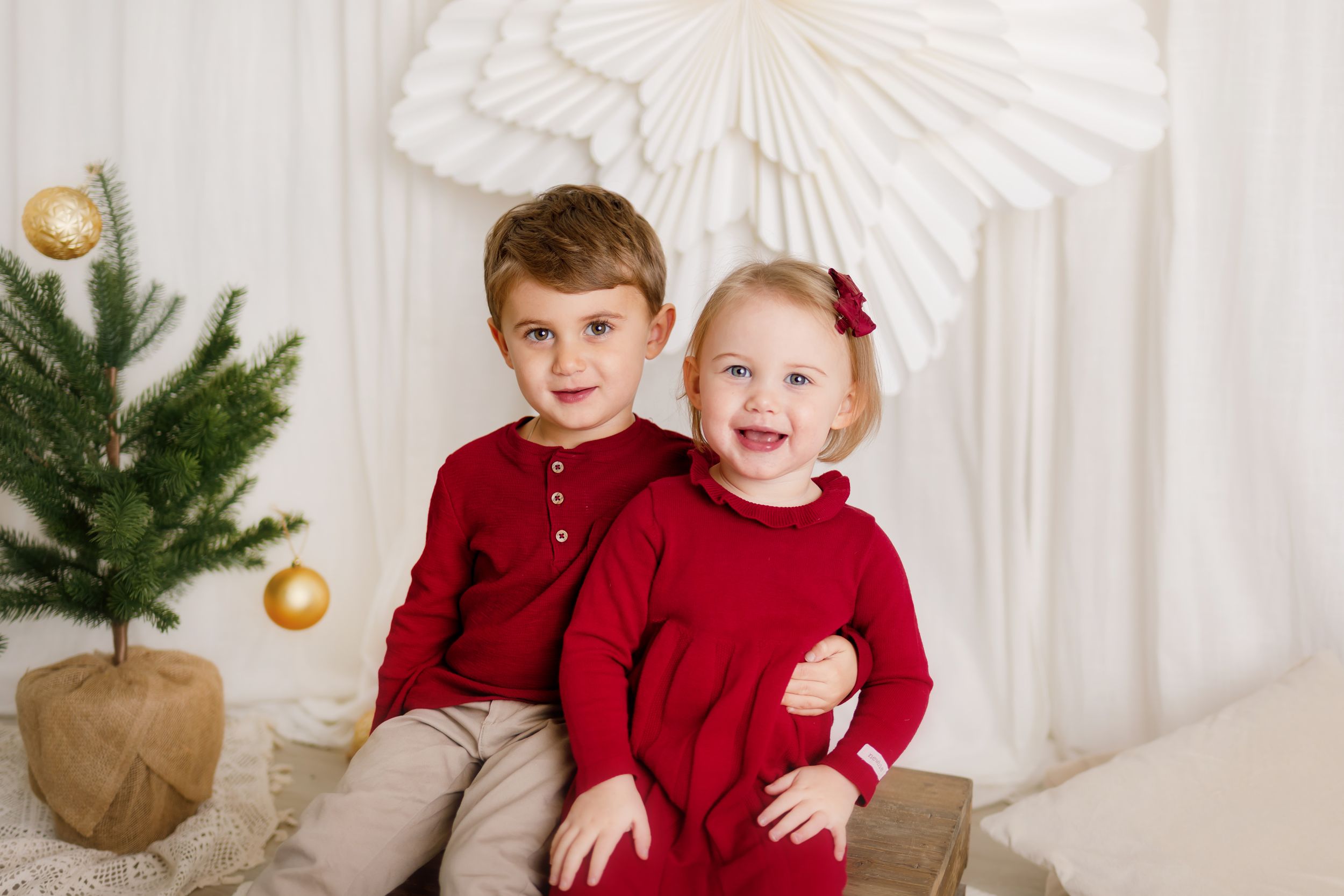 Two young siblings in matching red outfits pose together near a small Christmas tree with gold ornaments.