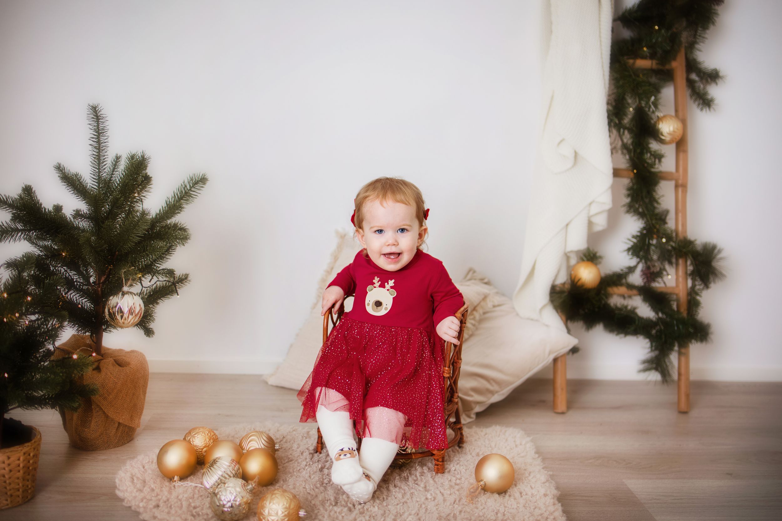 A child in a red velvet dress sits in front of a Christmas tree and gold ornament holiday display.
