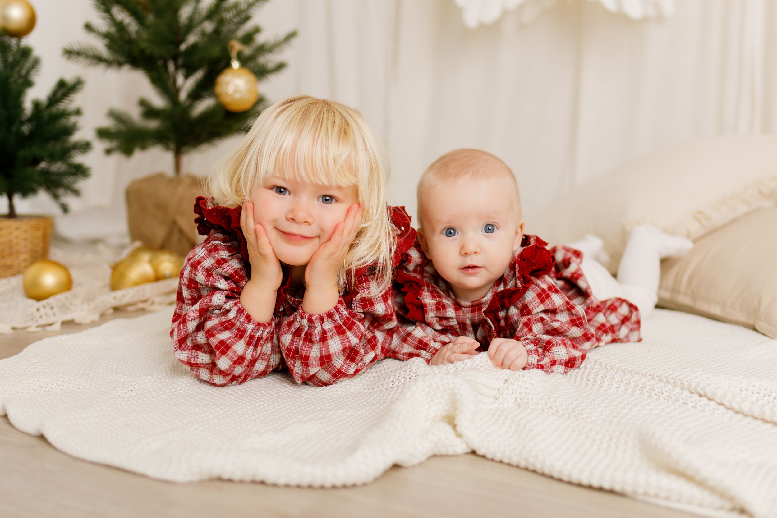 Two young siblings in matching red plaid Christmas outfits lying on a white blanket near a decorated holiday tree.