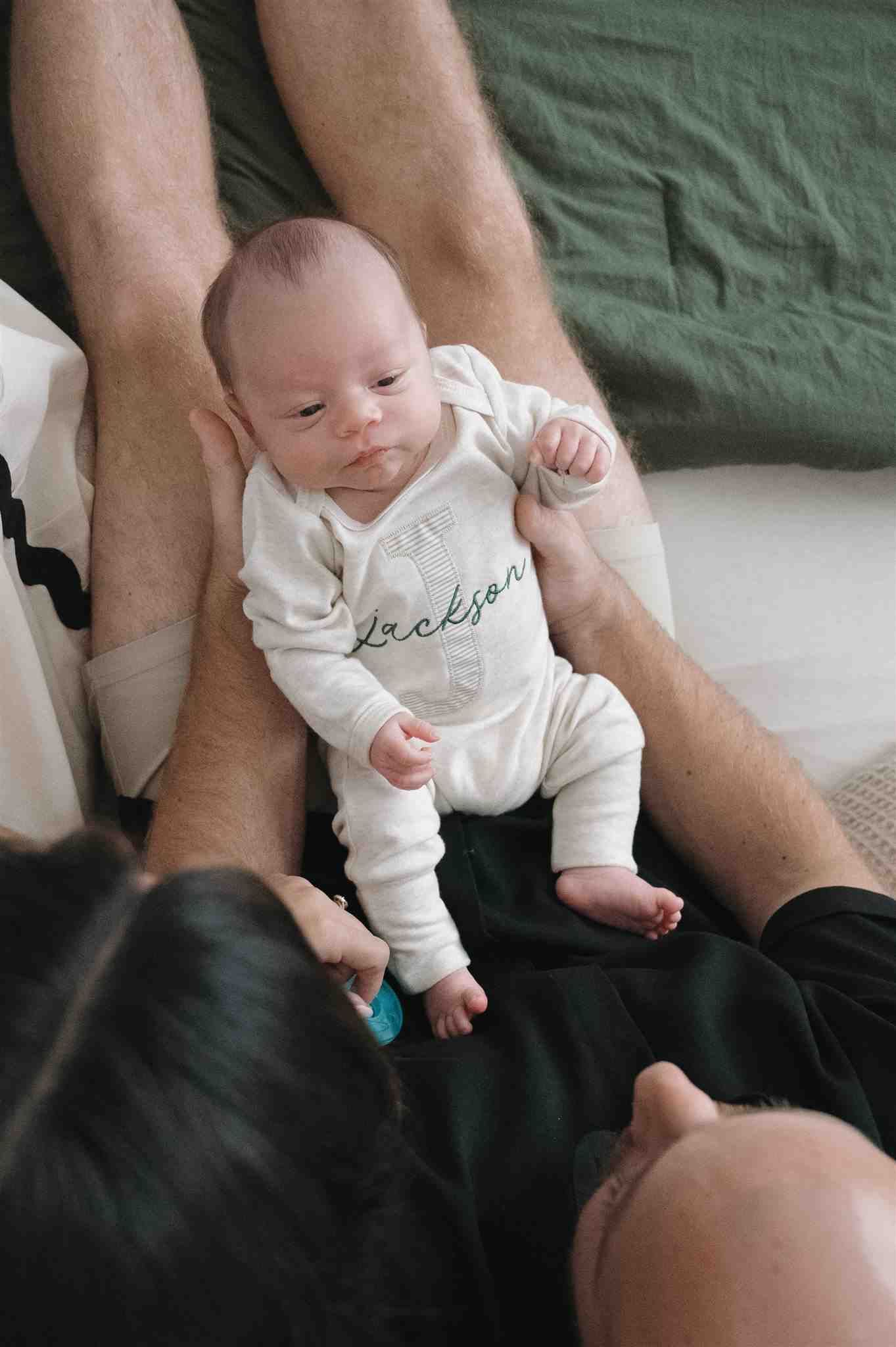 A small baby in white sleepwear rests against bare legs on a dark green couch.