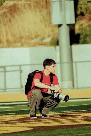 Photographer crouching on sports field with telephoto lens camera and backpack, wearing red shirt.