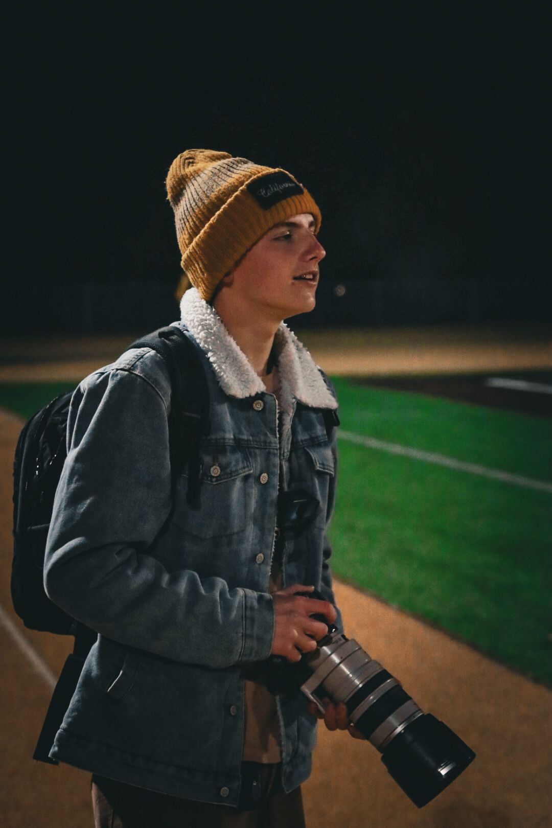 Photographer wearing a beanie and denim jacket holds a camera while shooting outdoors.