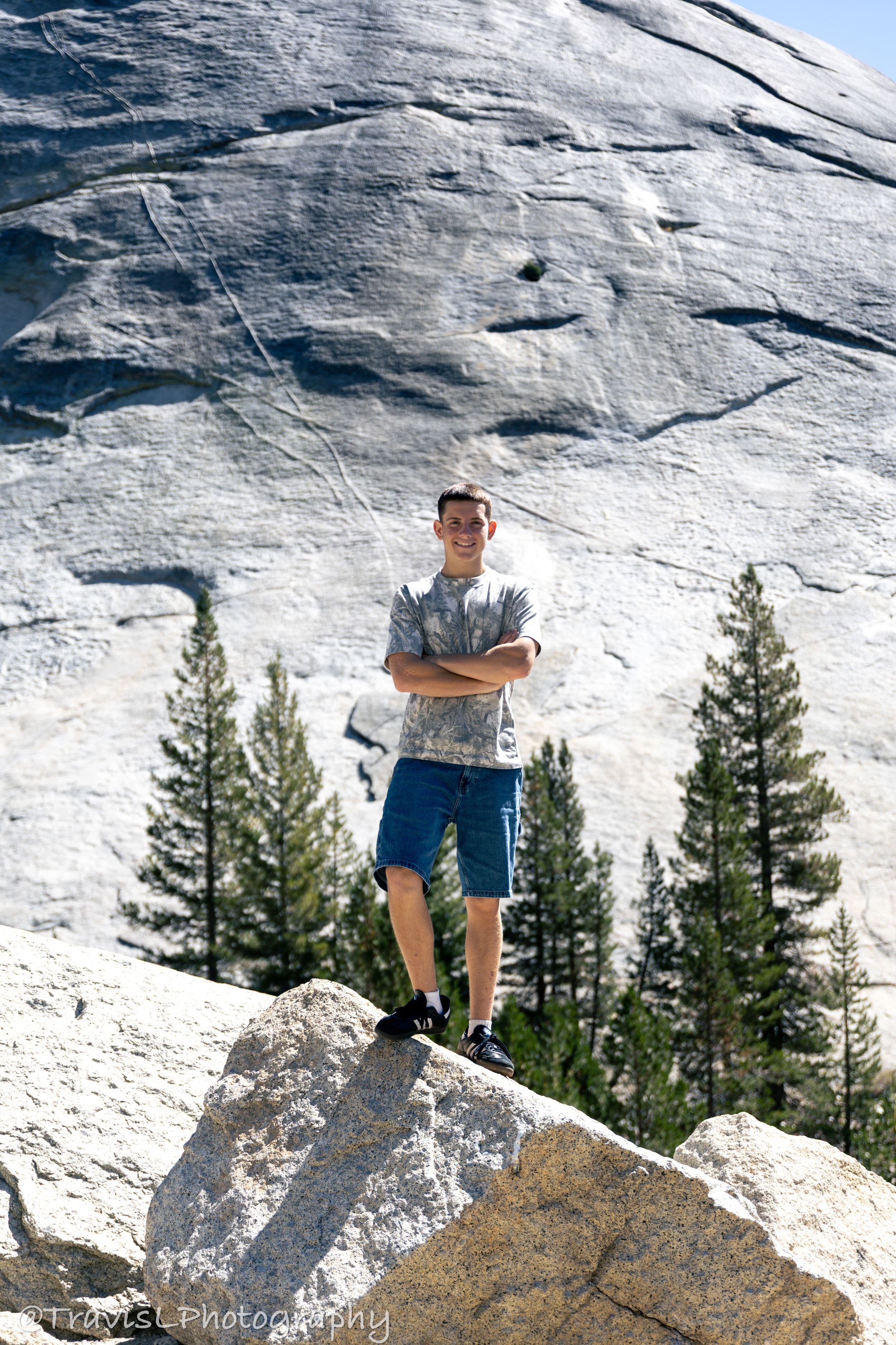 Person standing on rocky outcrop against white granite cliff face with pine trees below.