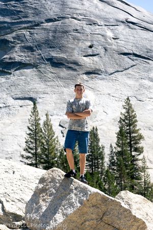 Person standing on rocky outcrop against white granite cliff face with pine trees below.