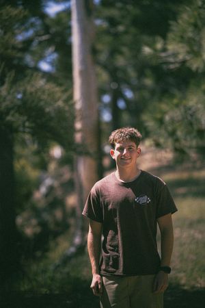 Young man in brown t-shirt smiling outdoors among green trees on a sunny day.