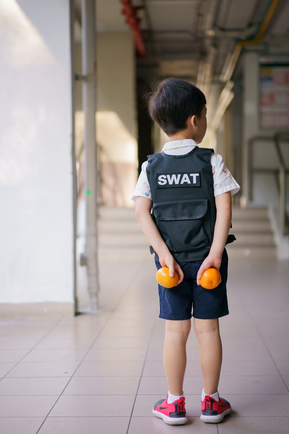 Child wearing SWAT vest standing in hallway holding oranges.