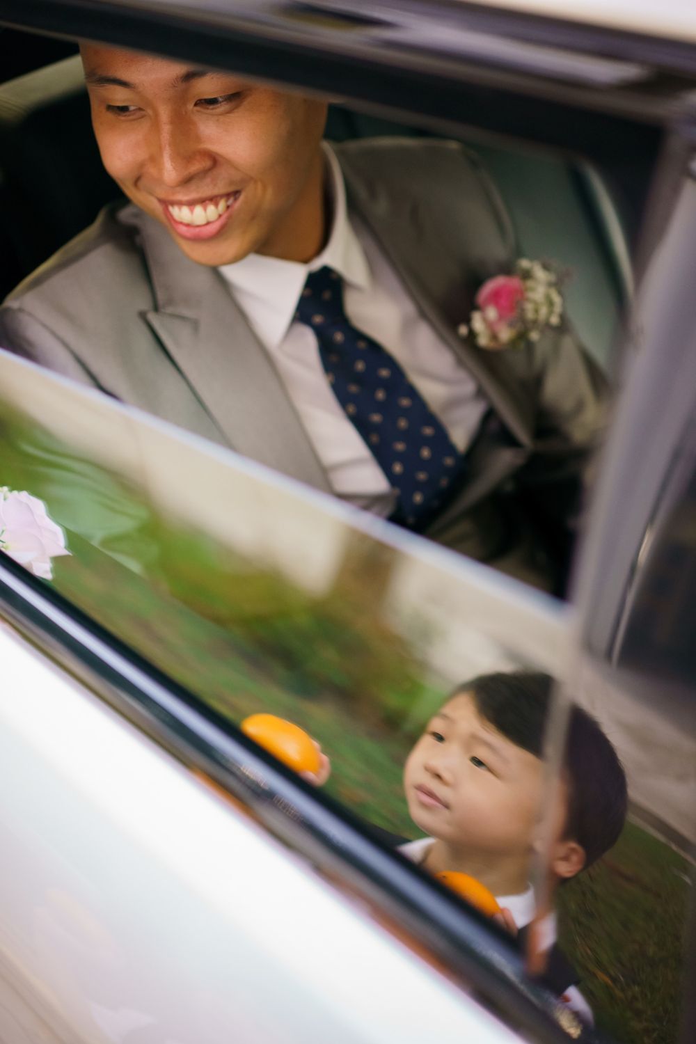 A joyful moment captured through a car window showing a groom and child on a wedding day.
