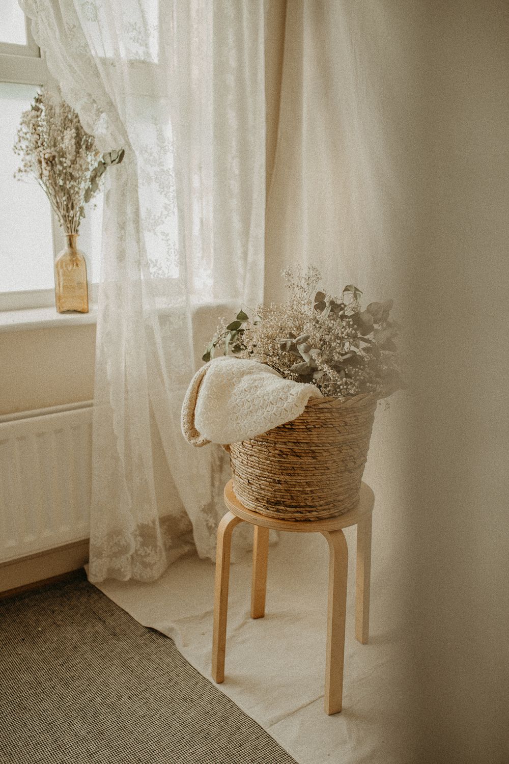 A cozy interior corner with sheer curtains, wooden stool, and dried flowers in a rustic rope-wrapped basket. Maternity studio in Beckenham