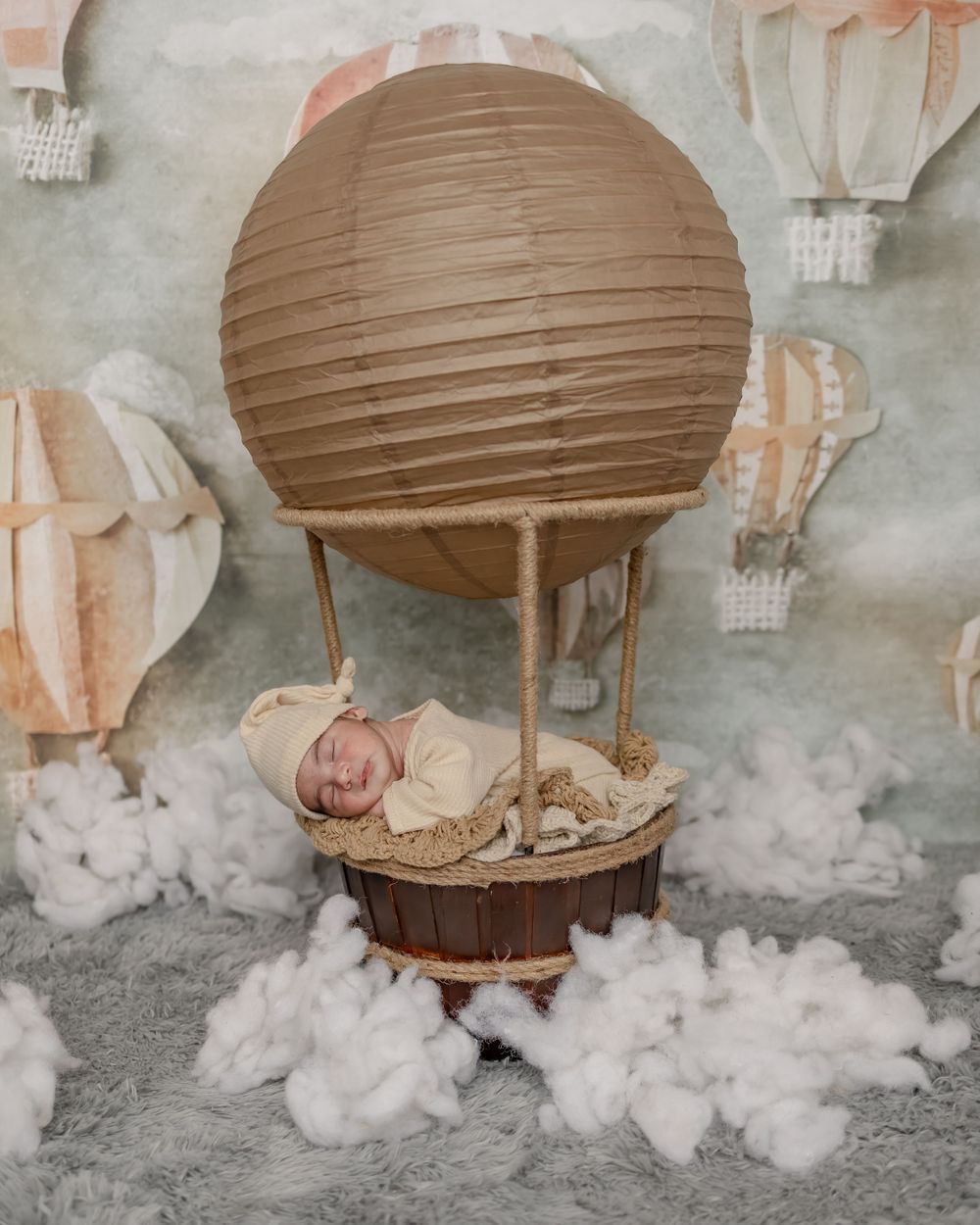 Newborn baby in vintage-style hot air balloon setup with clouds and dreamy backdrop.