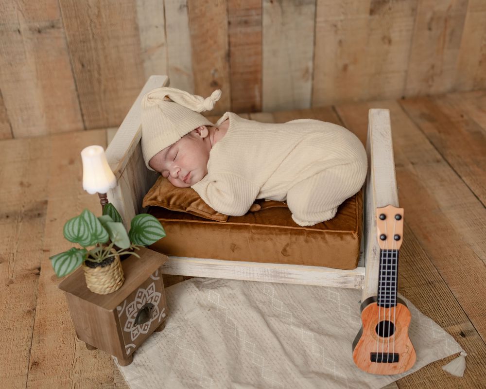 Sleeping newborn on wooden bed prop with ukulele and plant decor in rustic setting.