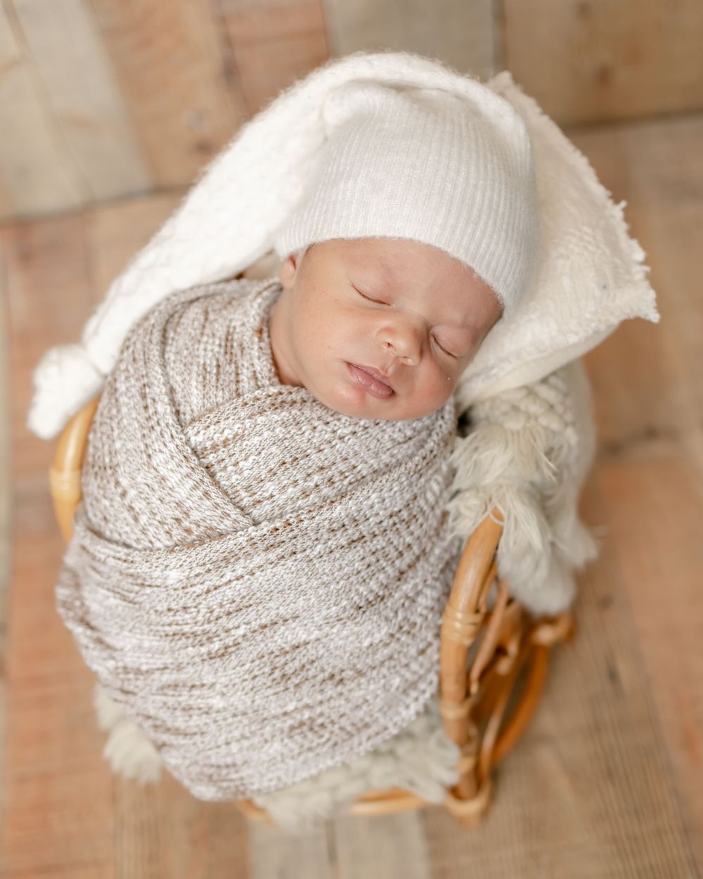 Sleeping newborn wrapped in knit blanket wearing white hat in wooden basket.