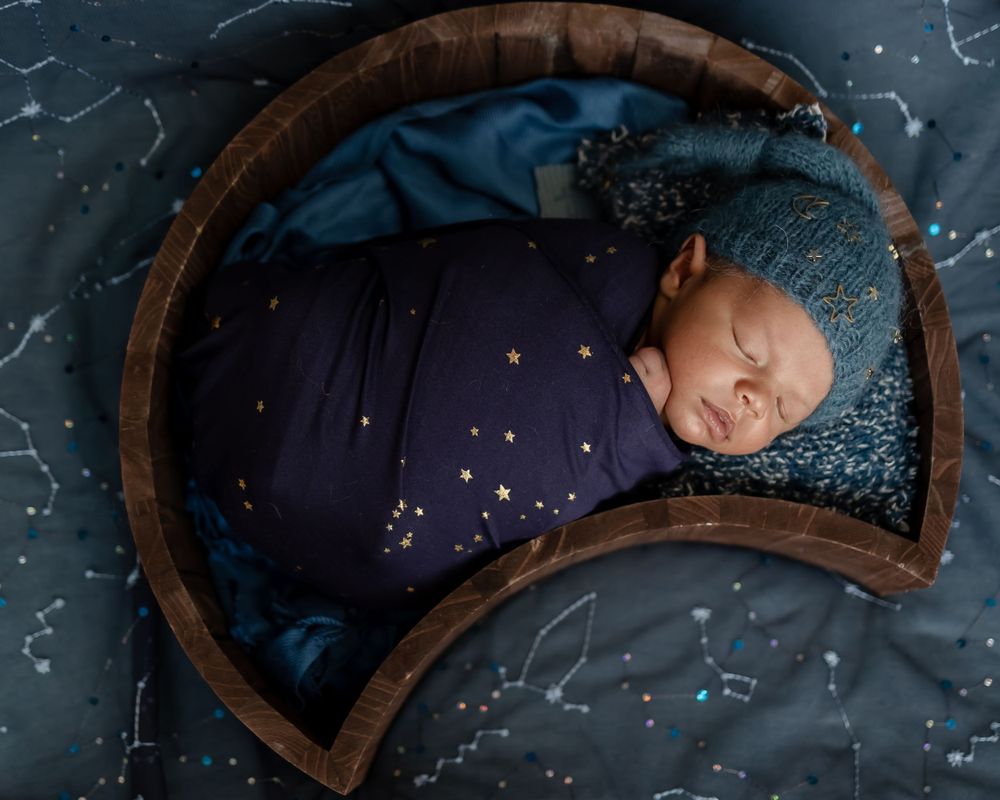 A newborn baby sleeps peacefully in a dark blue wrap inside a rustic wooden bowl against a starry backdrop.