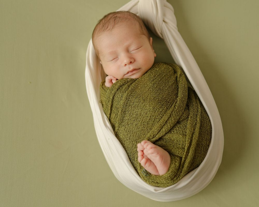 A sleeping newborn wrapped in olive green lies in a white fabric hammock.