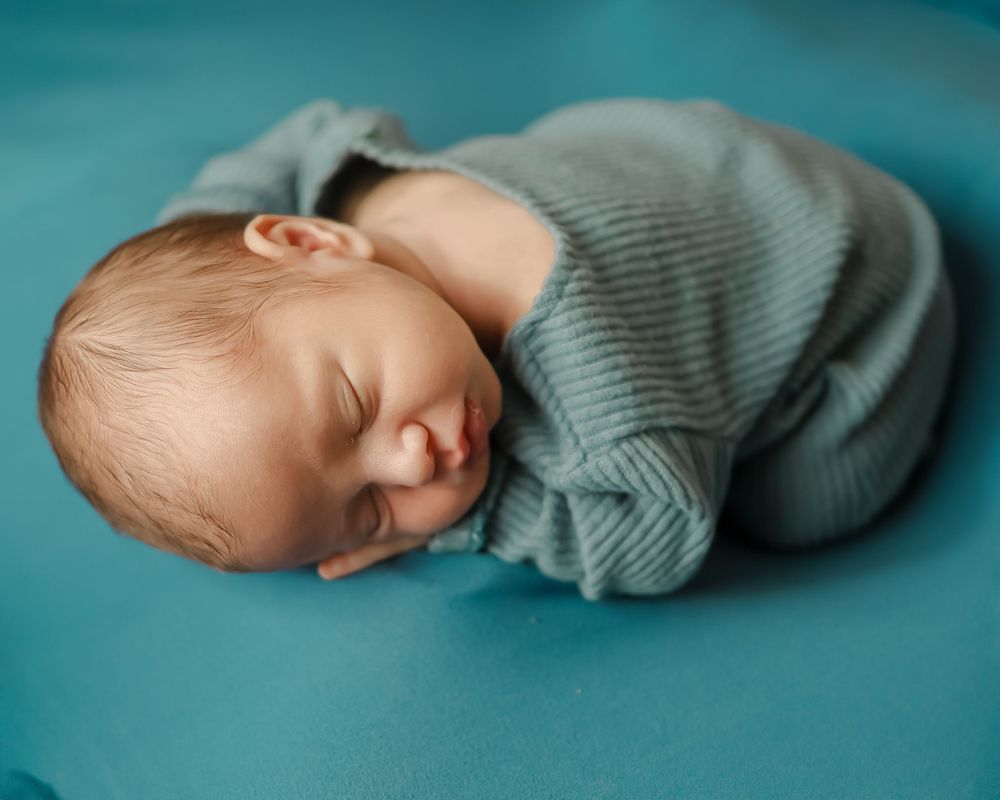 A peaceful sleeping newborn in soft teal clothing against a turquoise background.