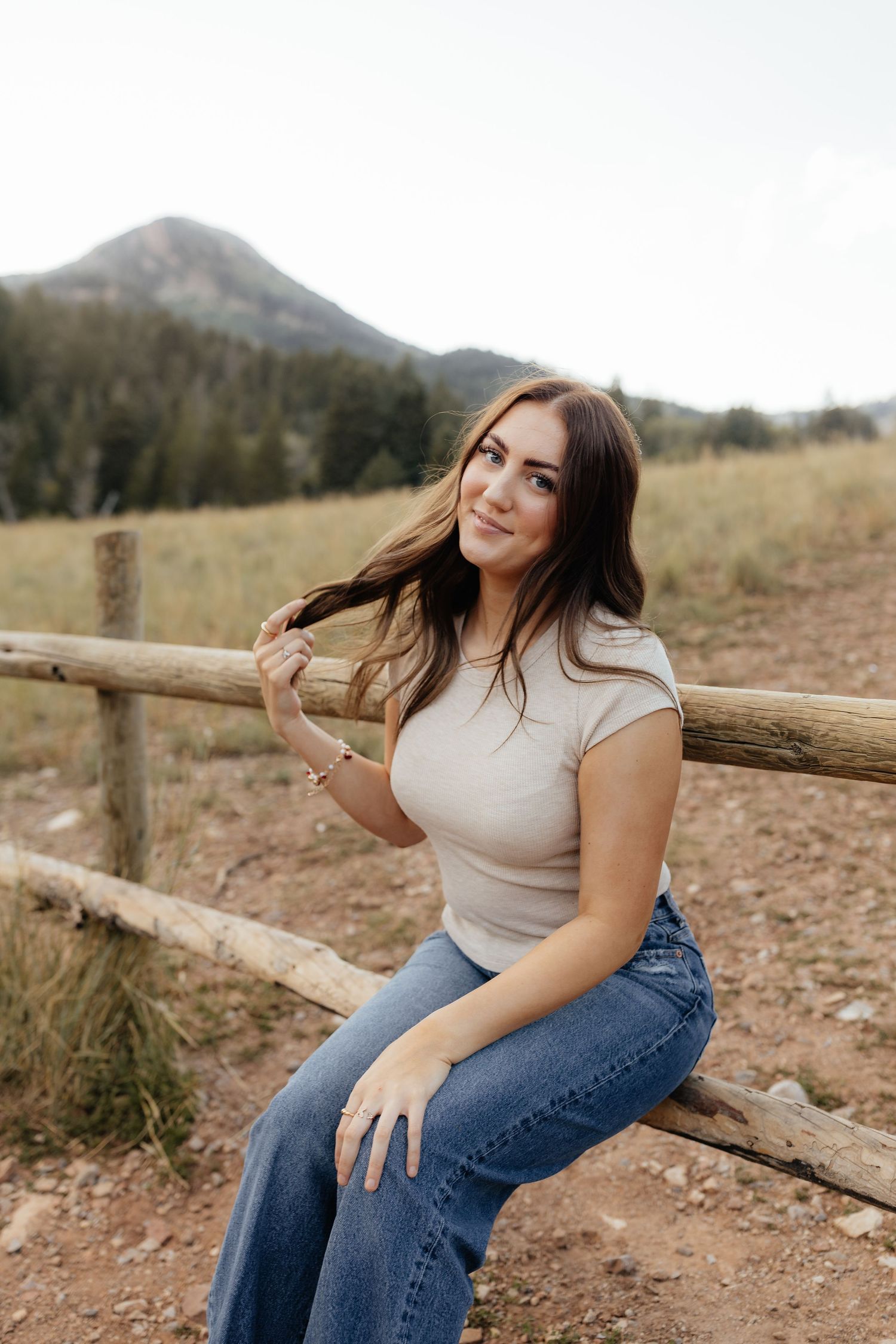 Brunette woman sits on a wooden fence in a mountain meadow with peaks in the background.
