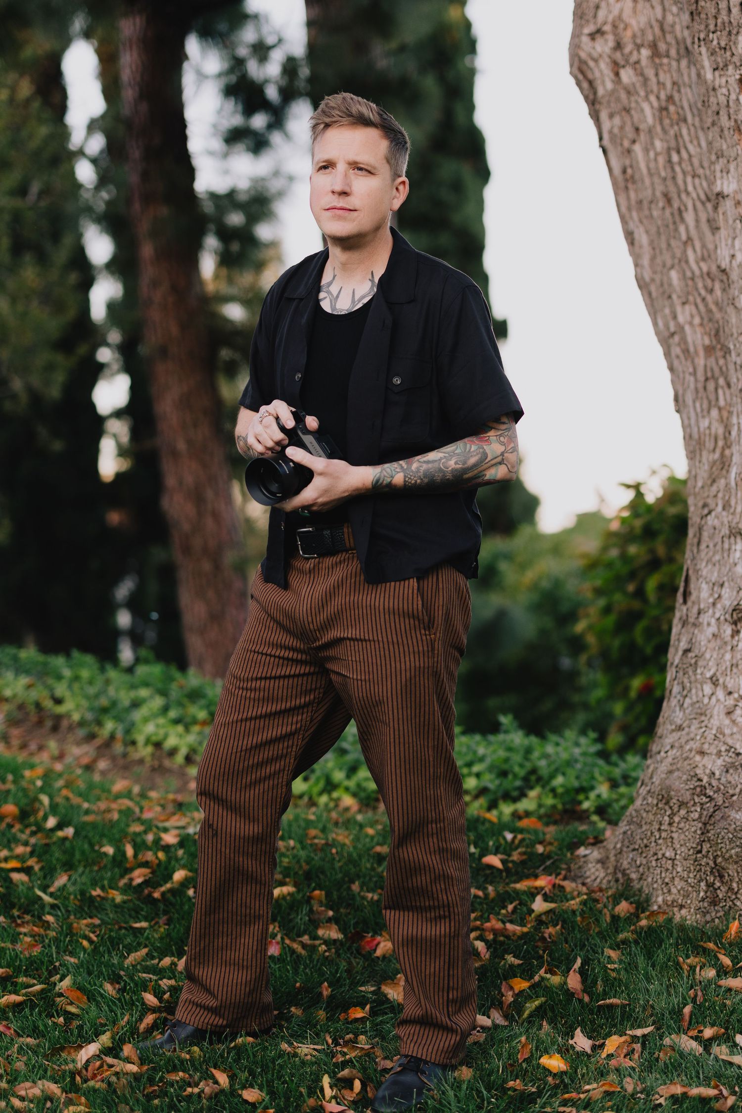 Tattooed male photographer holding camera standing among trees with autumn leaves on ground.