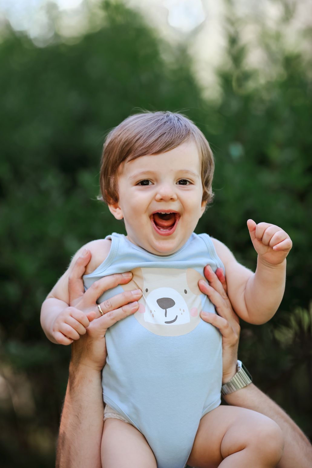 A joyful baby wearing a light blue onesie with a bear design smiles while being held up.