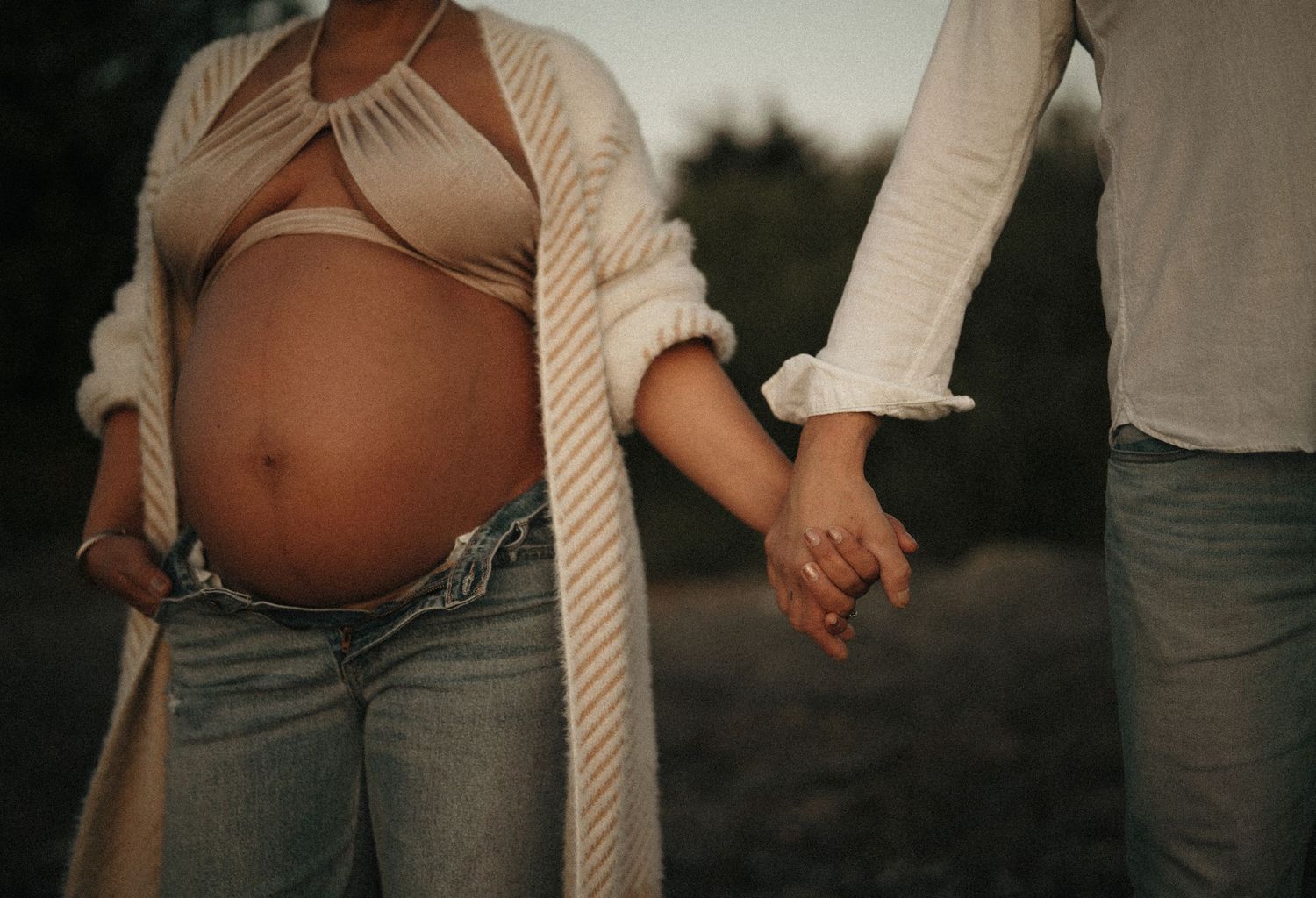 Couple holding hands with pregnant belly visible in casual beachwear.