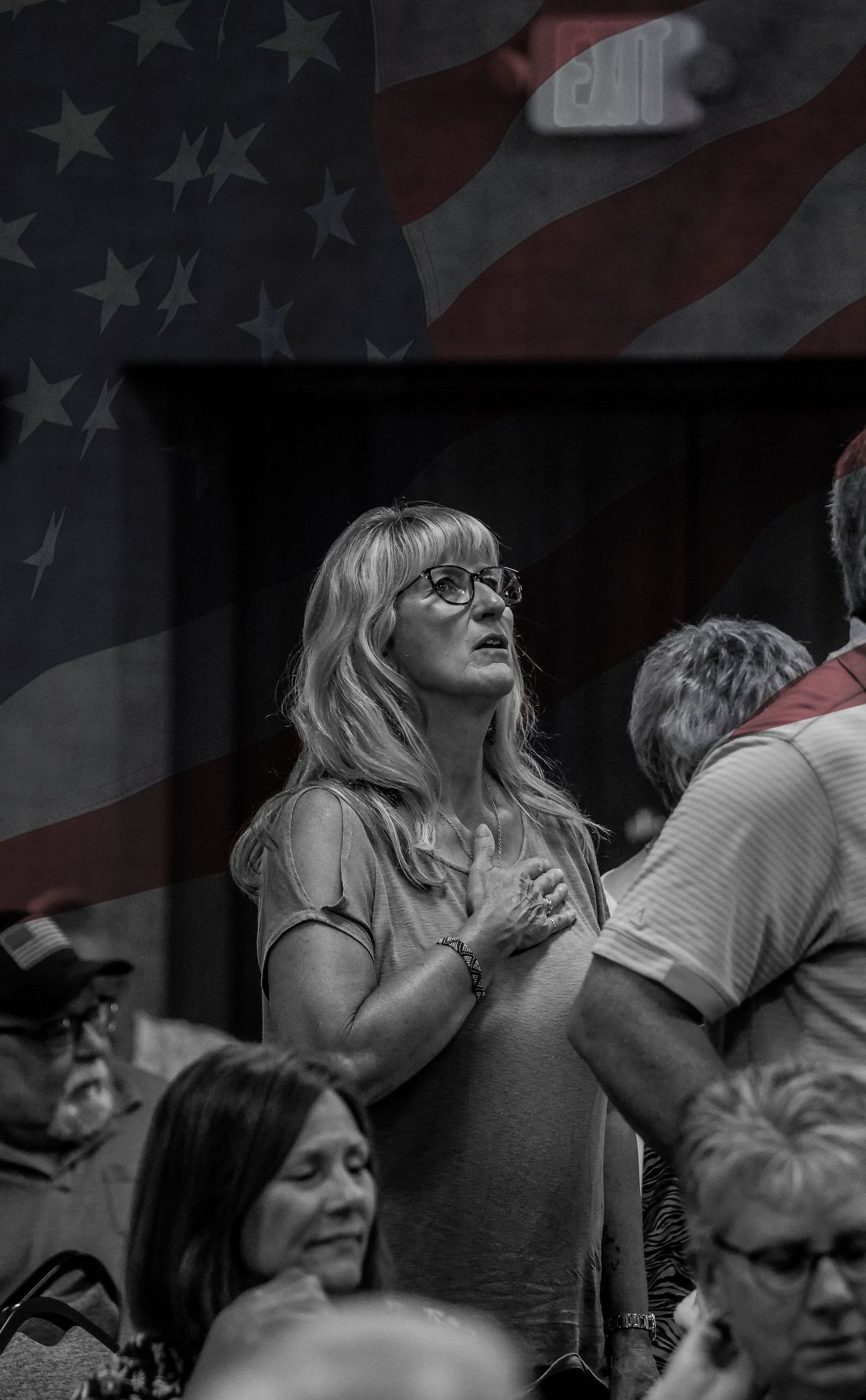 Person with glasses at patriotic gathering with American flag overlay in black and white.