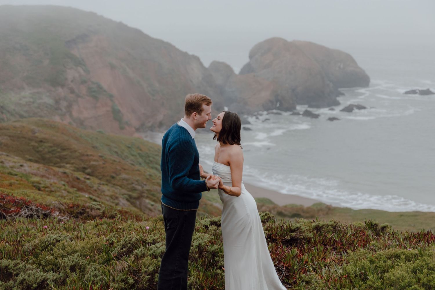 Foggy coastal cliffs create dramatic backdrop for intimate moment.