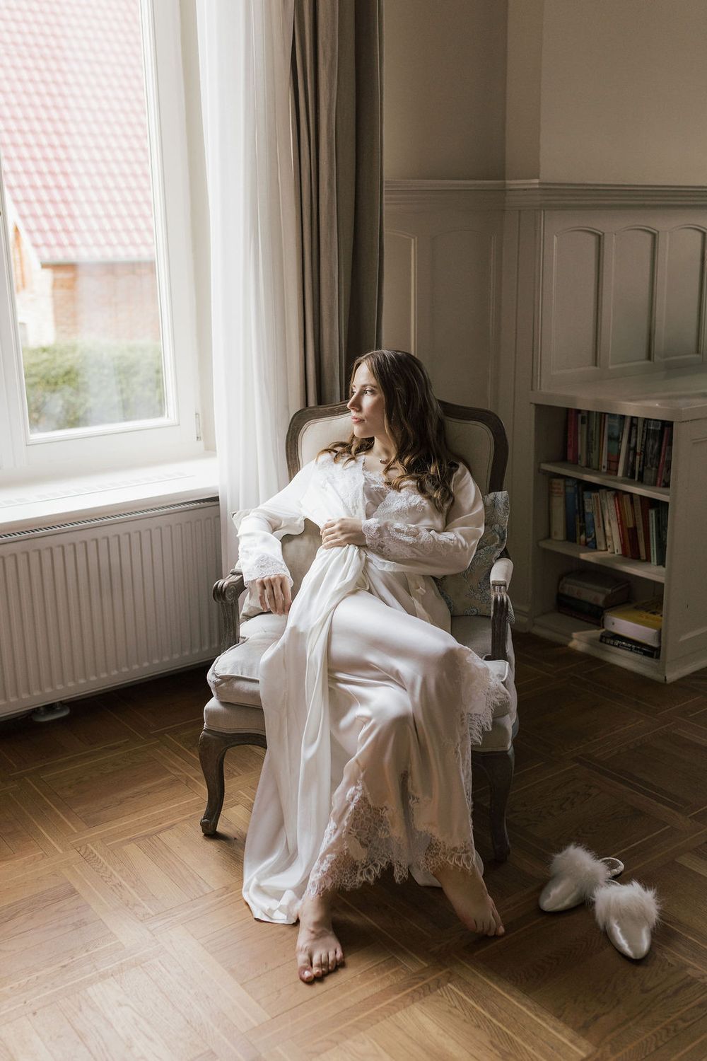 A figure in a flowing white dress sits elegantly in a vintage armchair by a window in a minimalist room.
