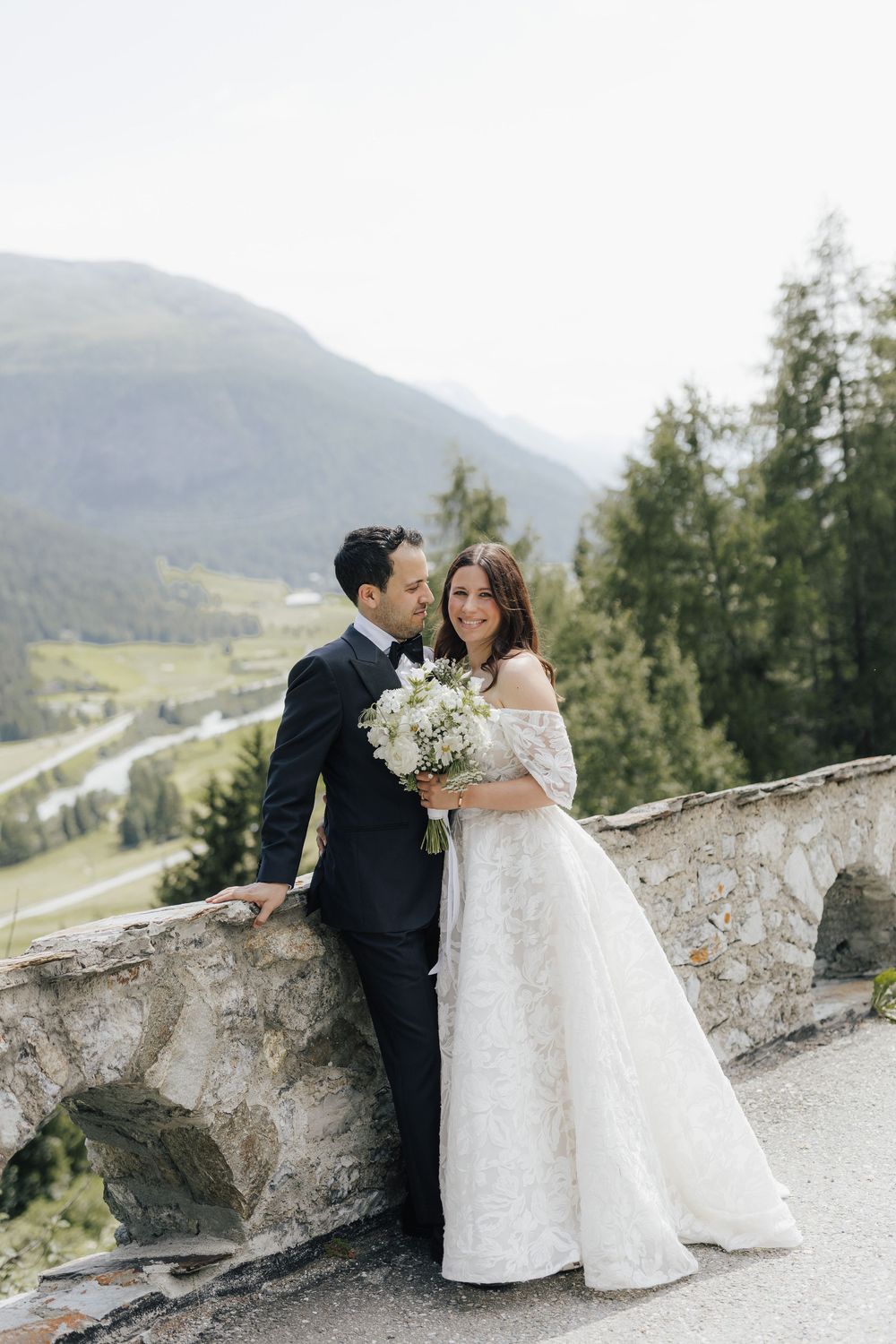 A couple in wedding attire shares romantic moments along a stone wall overlooking a scenic mountain landscape.