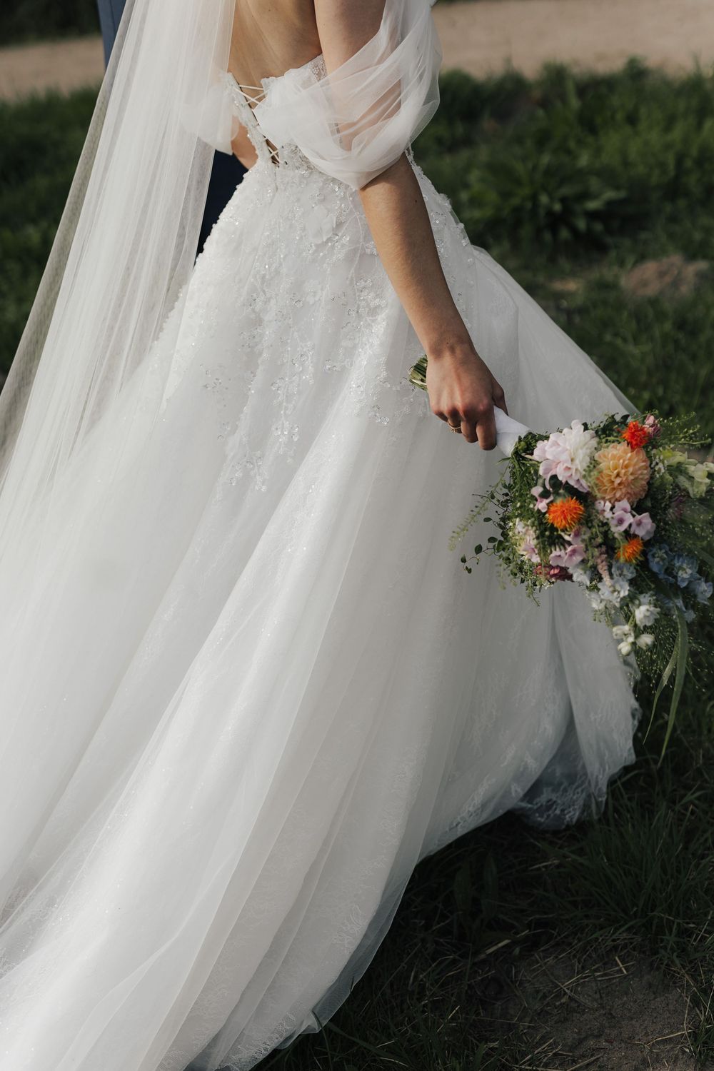 A bride in a flowing white wedding gown holds a vibrant wildflower bouquet against a natural outdoor backdrop.
