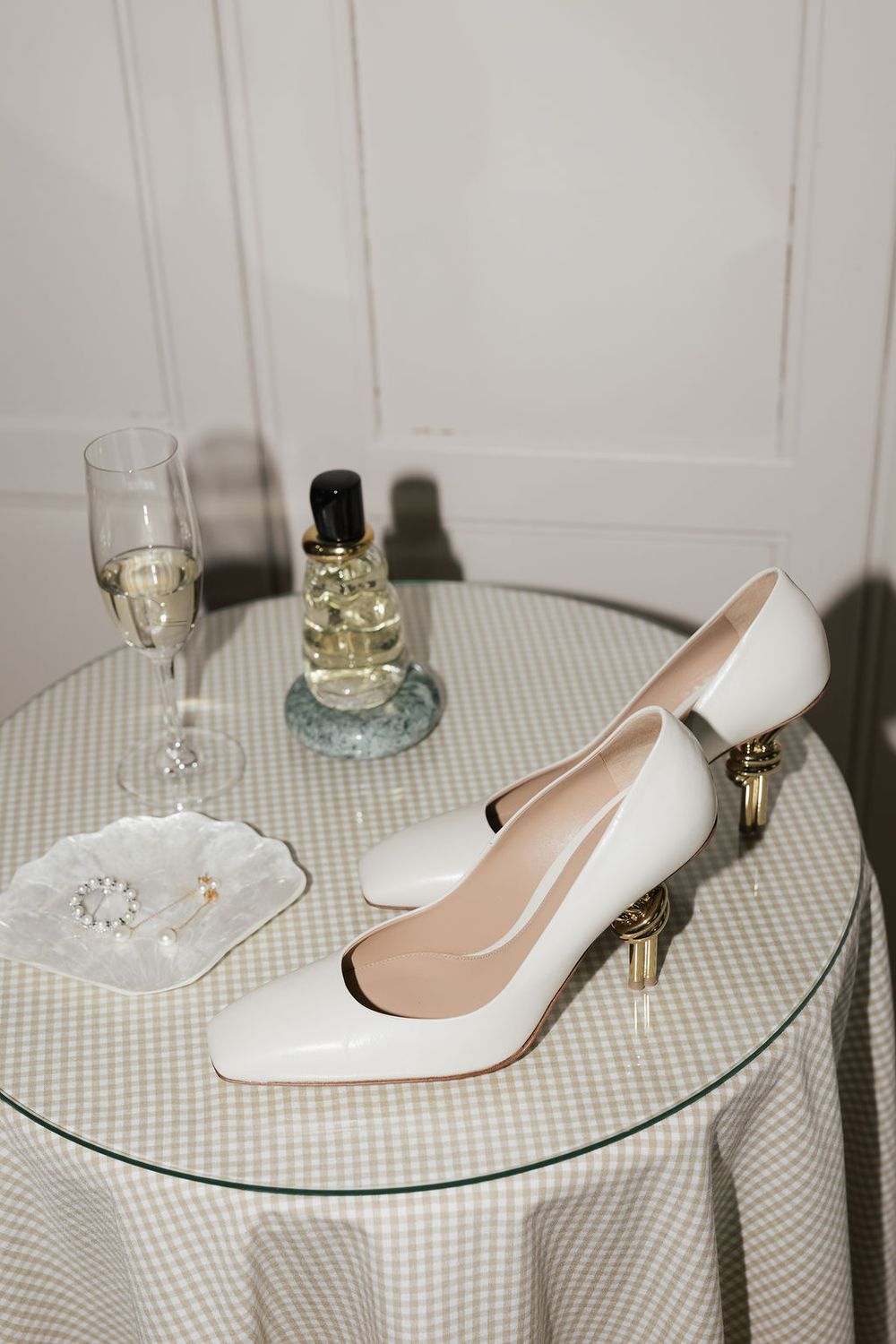 White high heel shoes displayed on a checkered tablecloth with wine glasses and perfume bottles in the background.