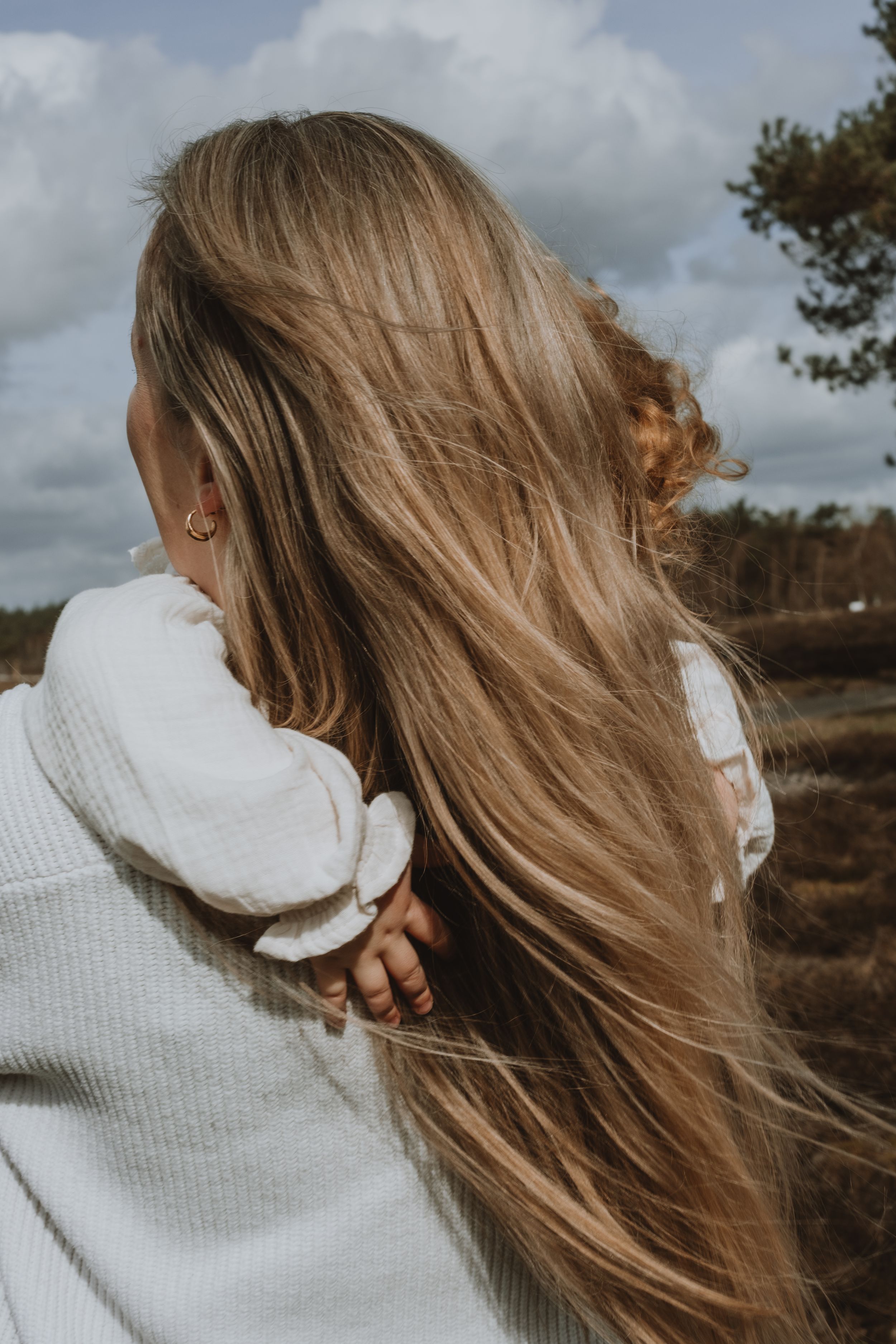 Back view of a woman with long flowing blonde hair holding a baby outdoors on a cloudy day.