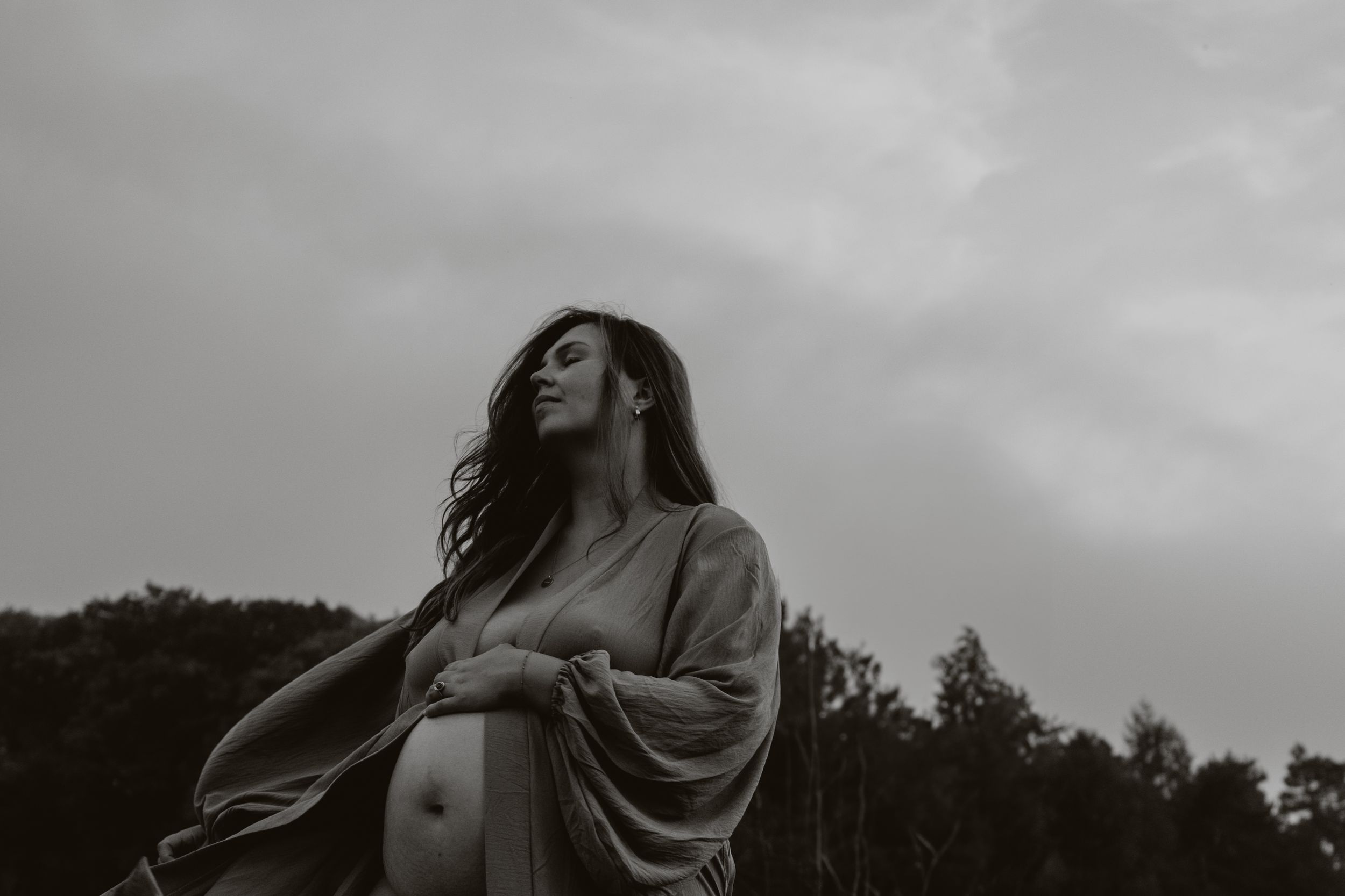 Pregnant woman in flowing robe stands outdoors under cloudy sky, cradling her baby bump in a moody black and white photo.