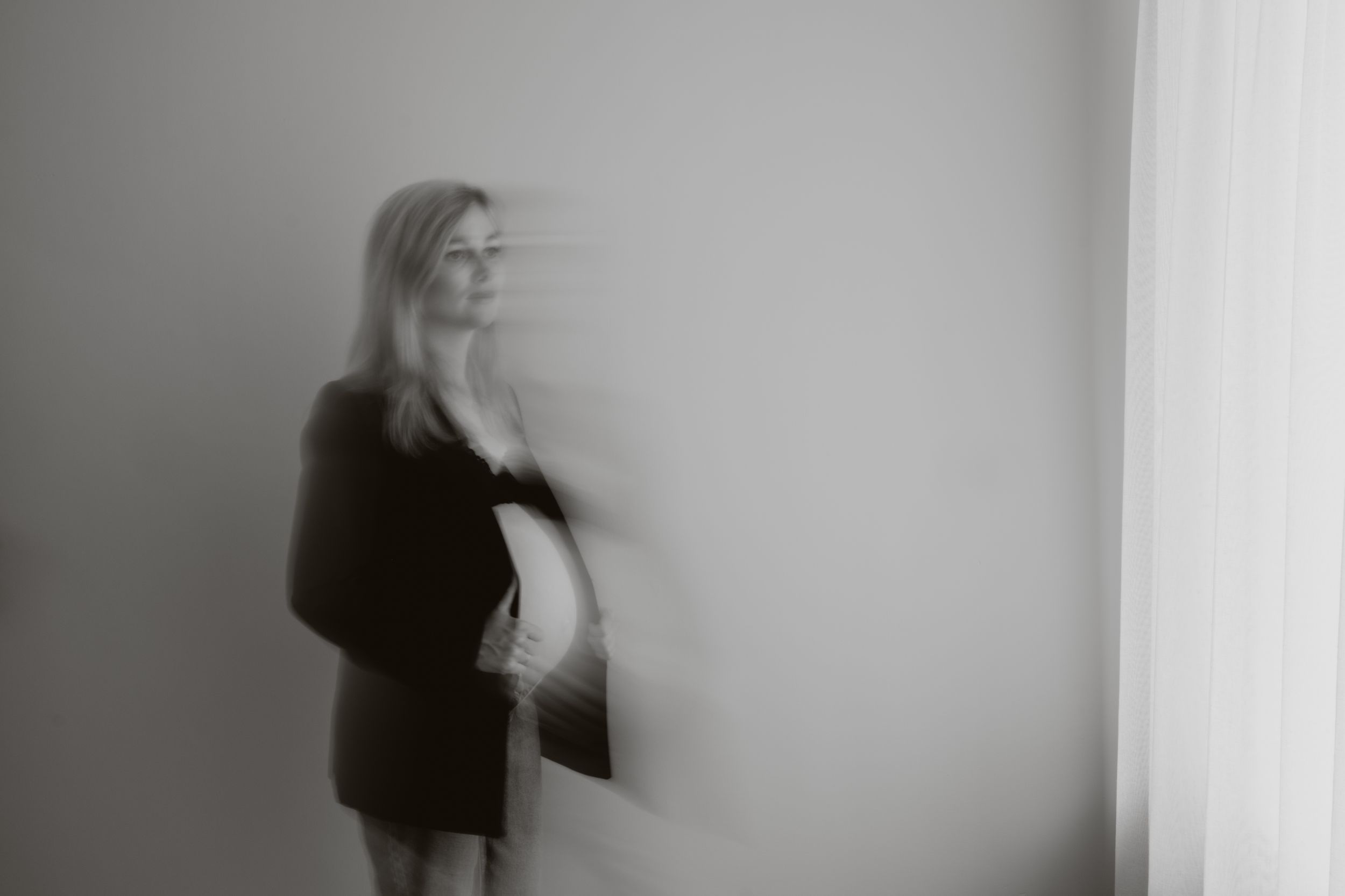 Blurred black and white long exposure photo of a pregnant woman standing near a window indoors.