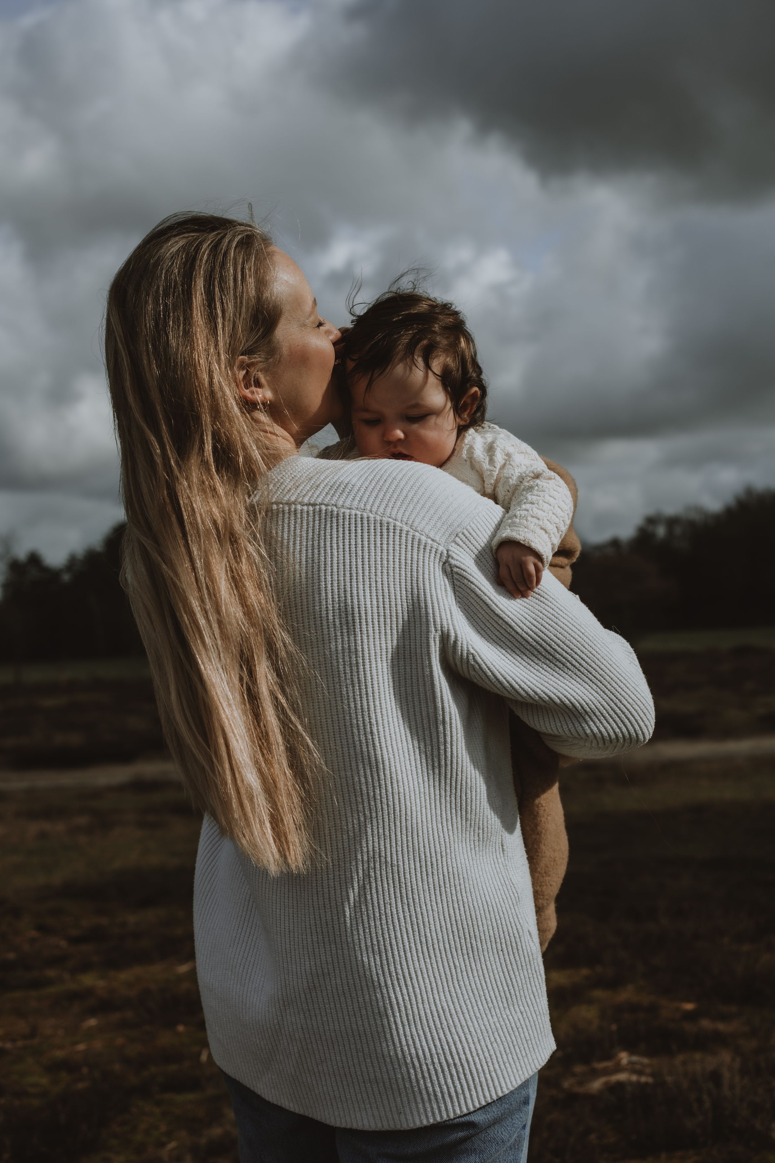 A mother with long blonde hair kisses her dark-haired baby outdoors under a cloudy sky.