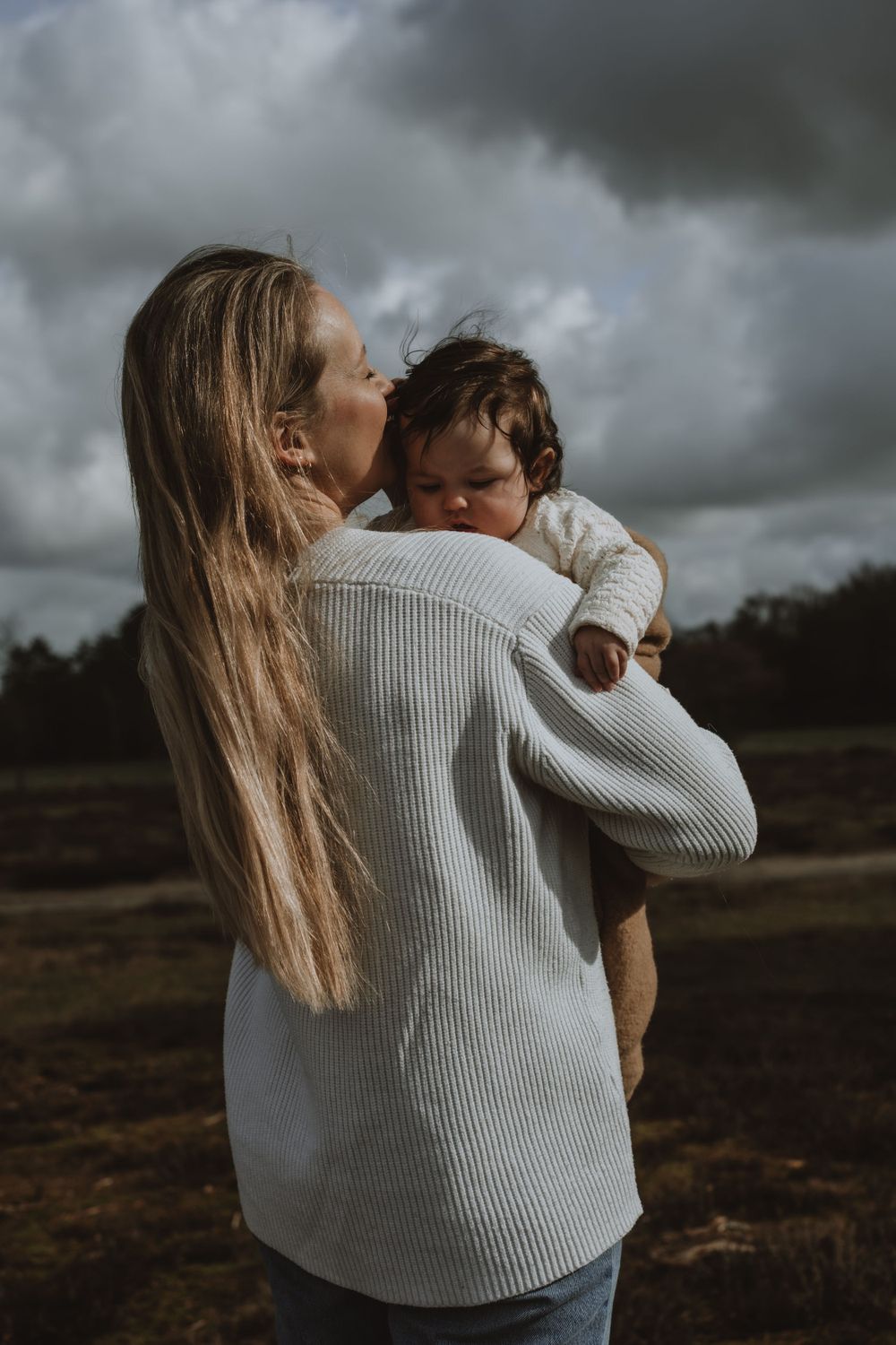 A mother with long blonde hair kisses her dark-haired baby outdoors under a cloudy sky.