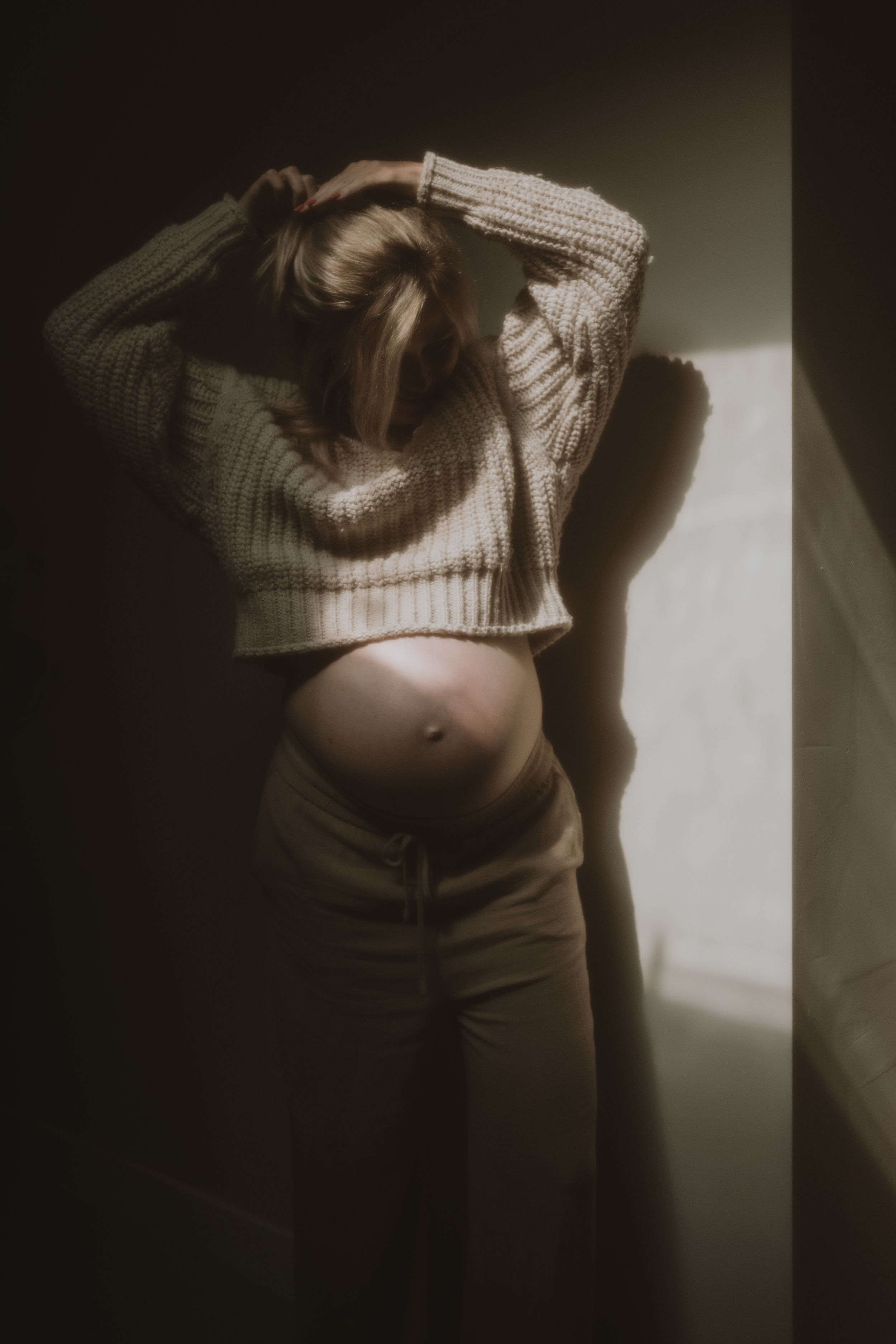 Pregnant woman in cream knit sweater poses near window, cradling bump with dramatic shadow on wall.
