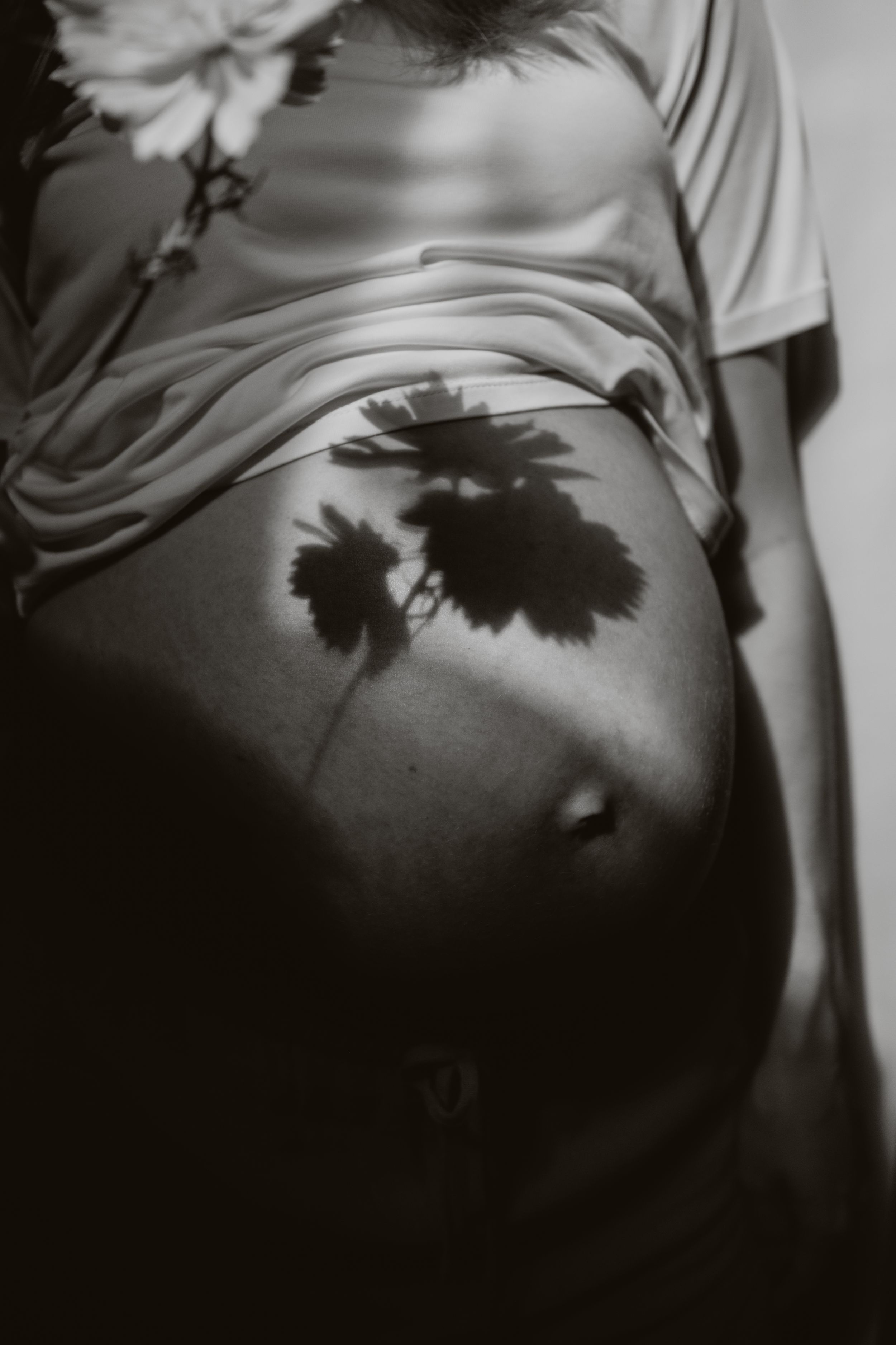 Black and white close-up of pregnant belly with flower shadows cast across white fabric top.