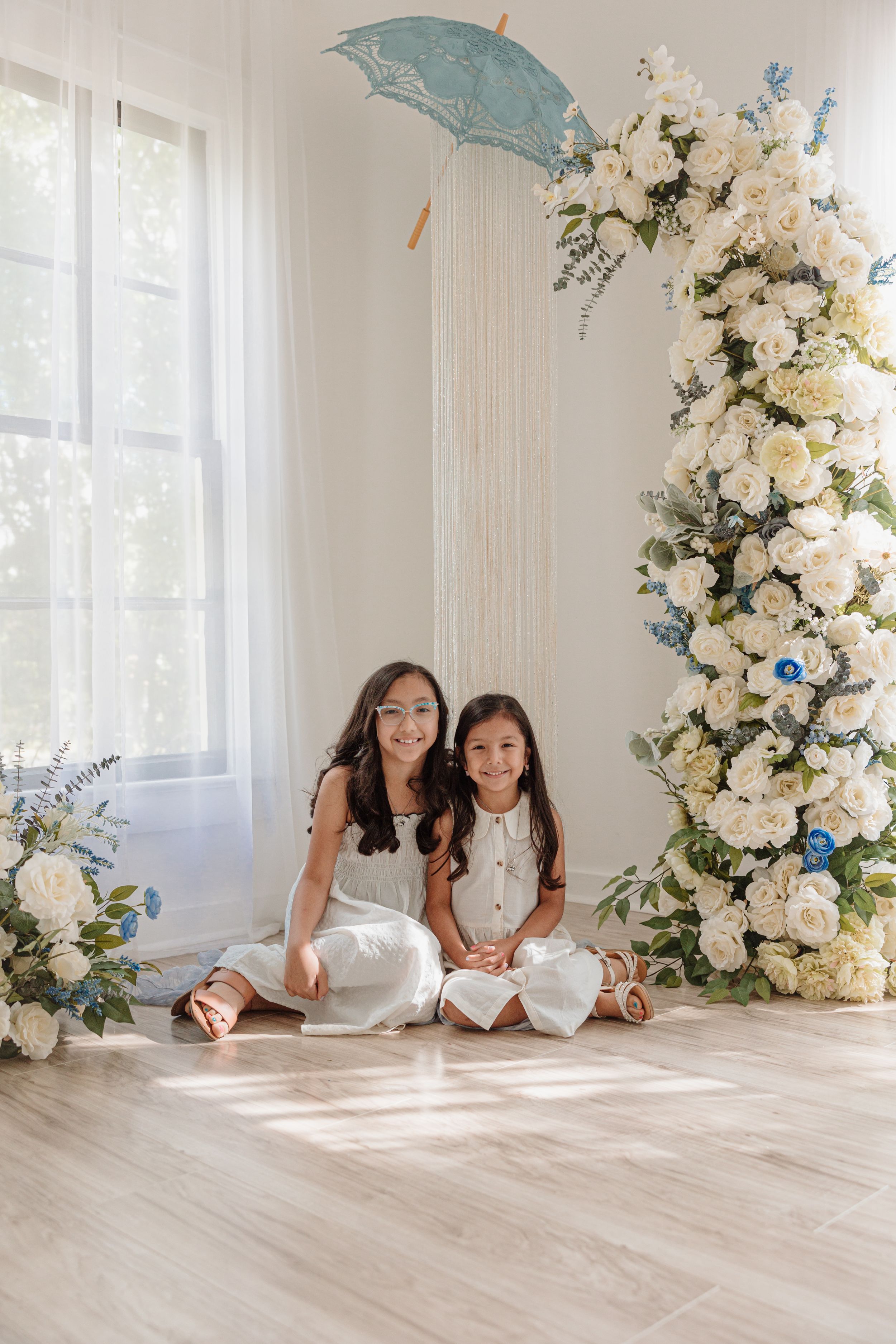 Two people sitting on floor by white floral arrangements and blue parasol decorations.