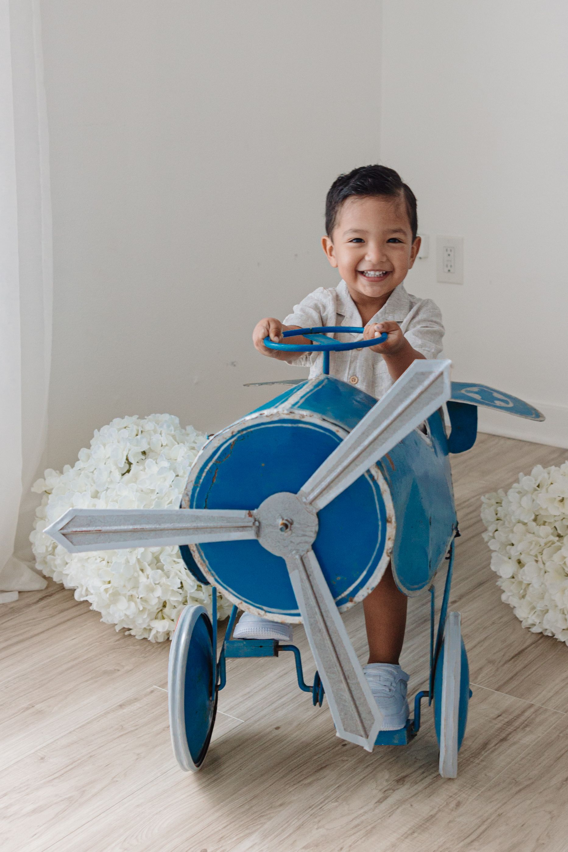 Child playing with blue toy airplane prop surrounded by white hydrangea arrangements.