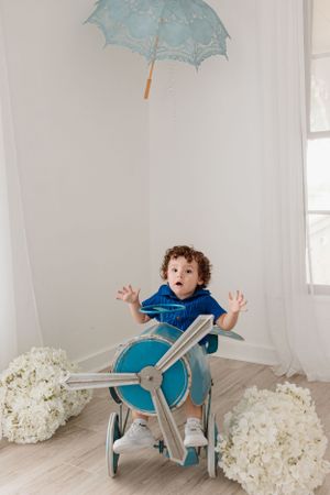 A small child sits in a vintage airplane prop with a blue parasol hanging overhead and white floral decorations.