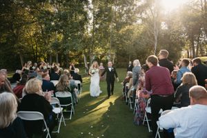 A beautiful outdoor wedding ceremony at sunset with guests seated on white chairs amid lush greenery.