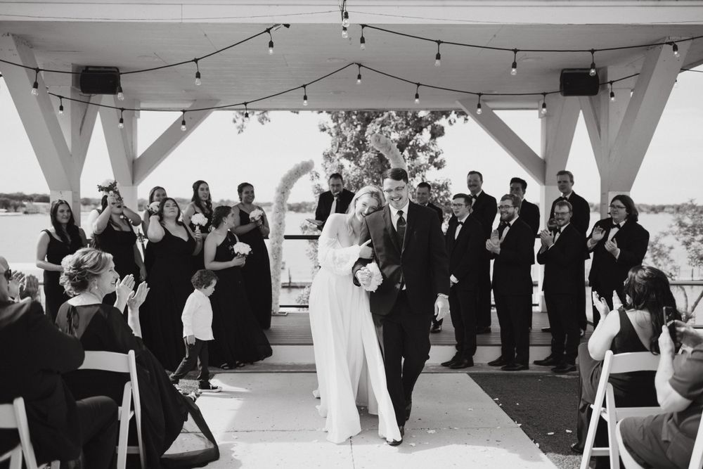 Black and white photo of newlyweds walking down aisle under reception pavilion with string lights.