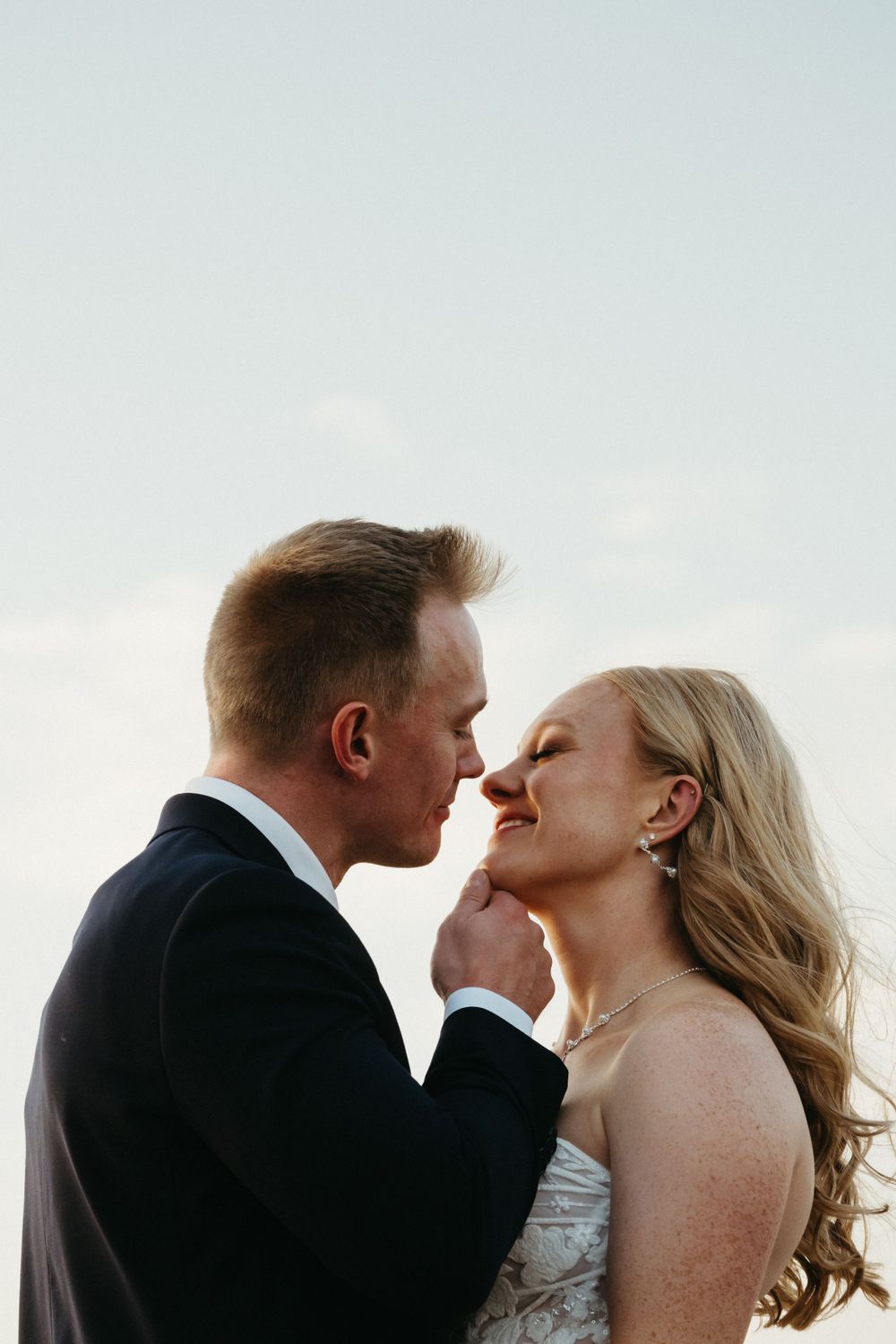 Romantic couple in wedding attire sharing an intimate moment against a bright sky.