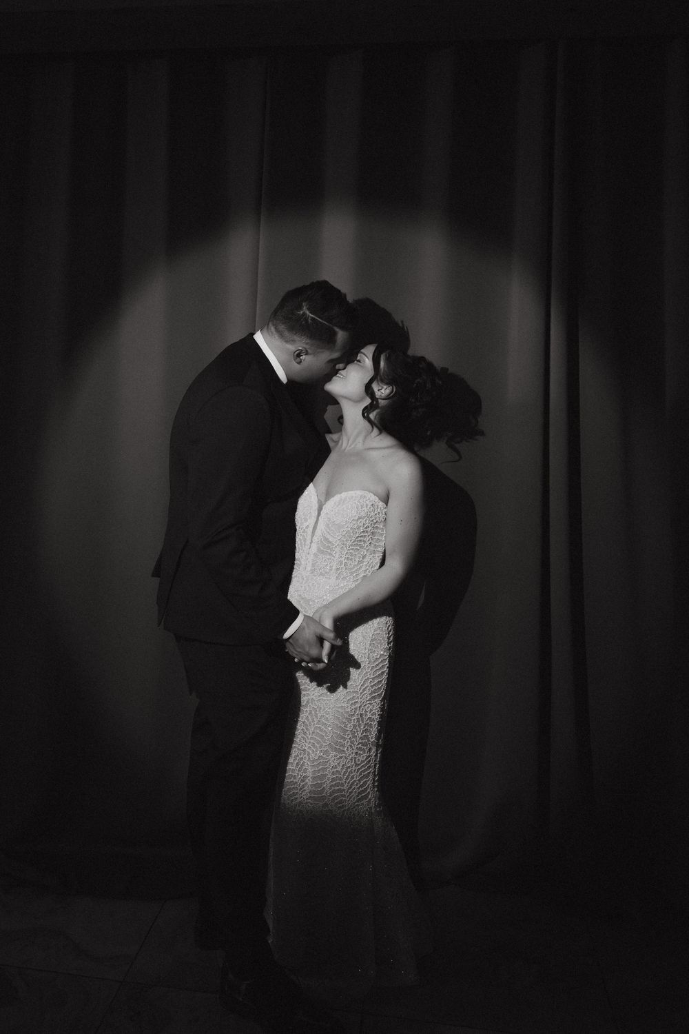 Black and white wedding photo of bride and groom kissing under spotlight against draped curtain backdrop.