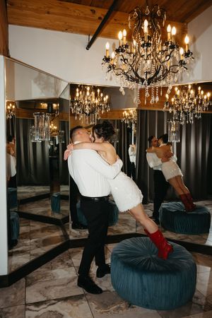 Couple sharing a romantic kiss in elegant room with crystal chandelier and mirrored walls, bride in white dress and red boots.