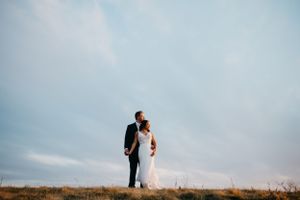 A couple stands together on a hilltop against a cloudy blue sky in wedding attire.
