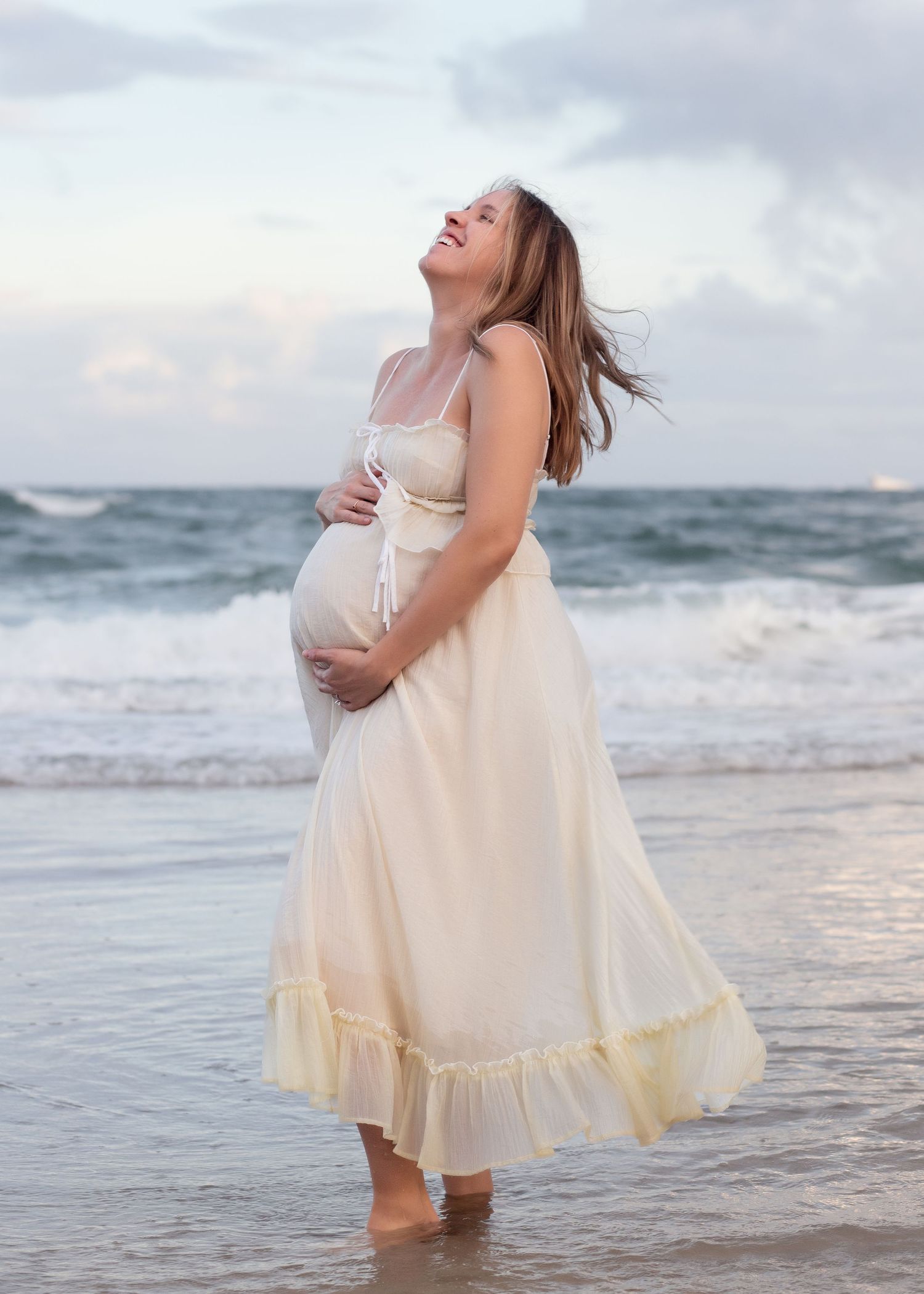 A person in a flowing pink dress poses on a beach at sunset with ocean waves in the background.